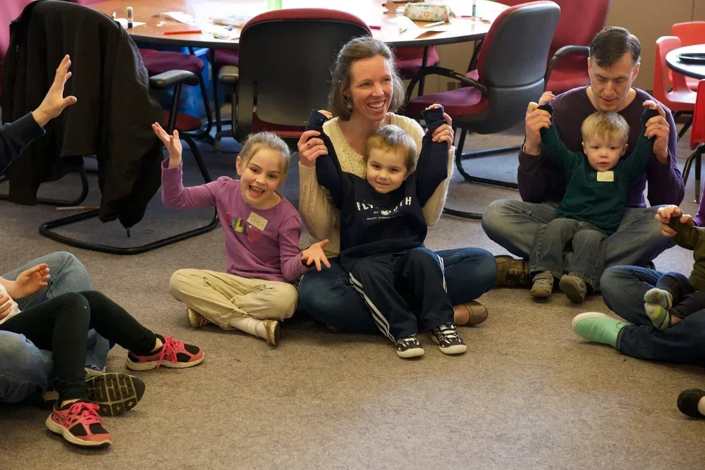 A group of children and adults sitting on the floor in a circle, smiling and raising their hands, in an indoor setting with chairs and tables around.