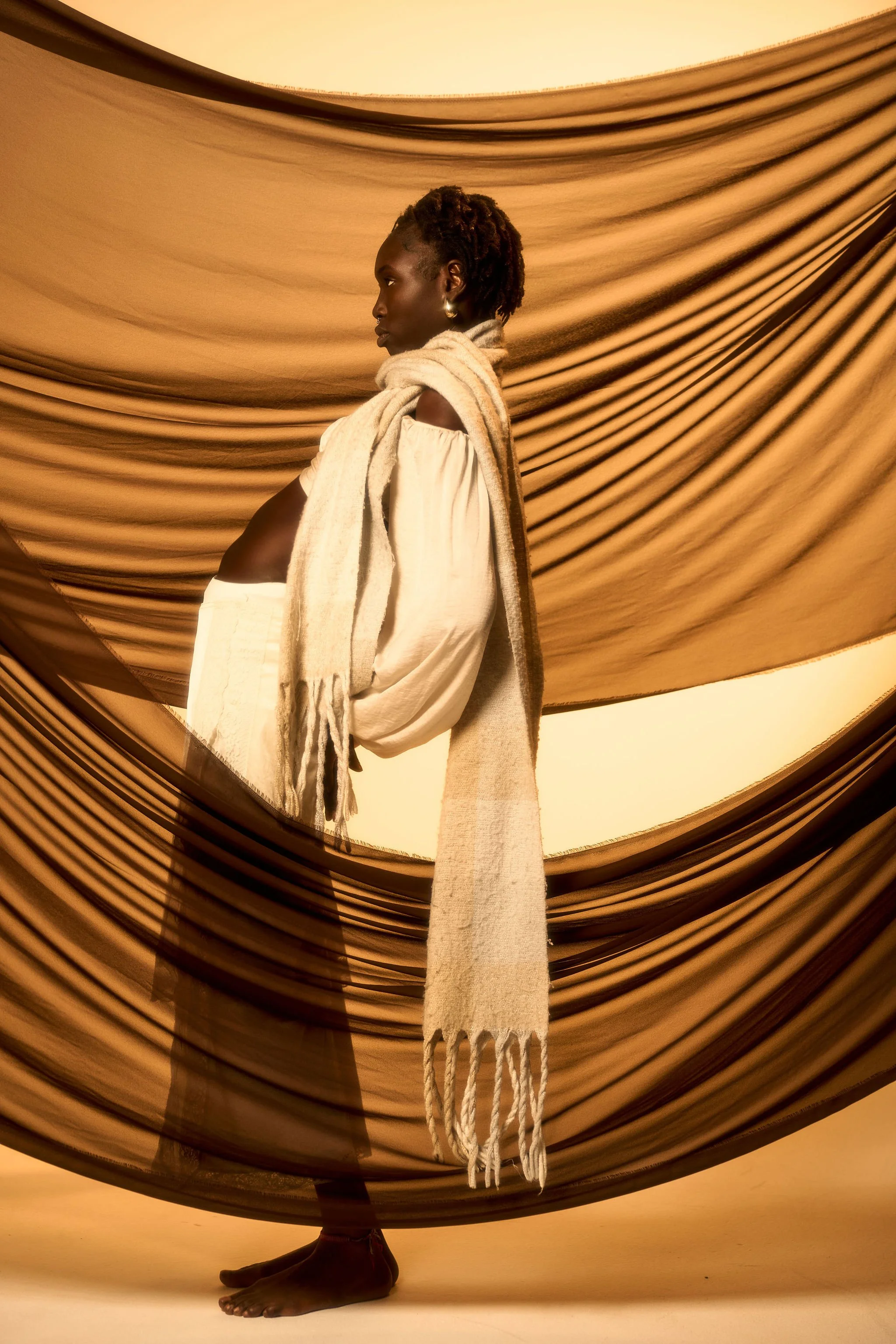 A woman with dark skin, dressed in white, stands with her eyes closed against fabric draped in the background, with a beige scarf wrapped around her shoulders and a long fringe scarf hanging down.