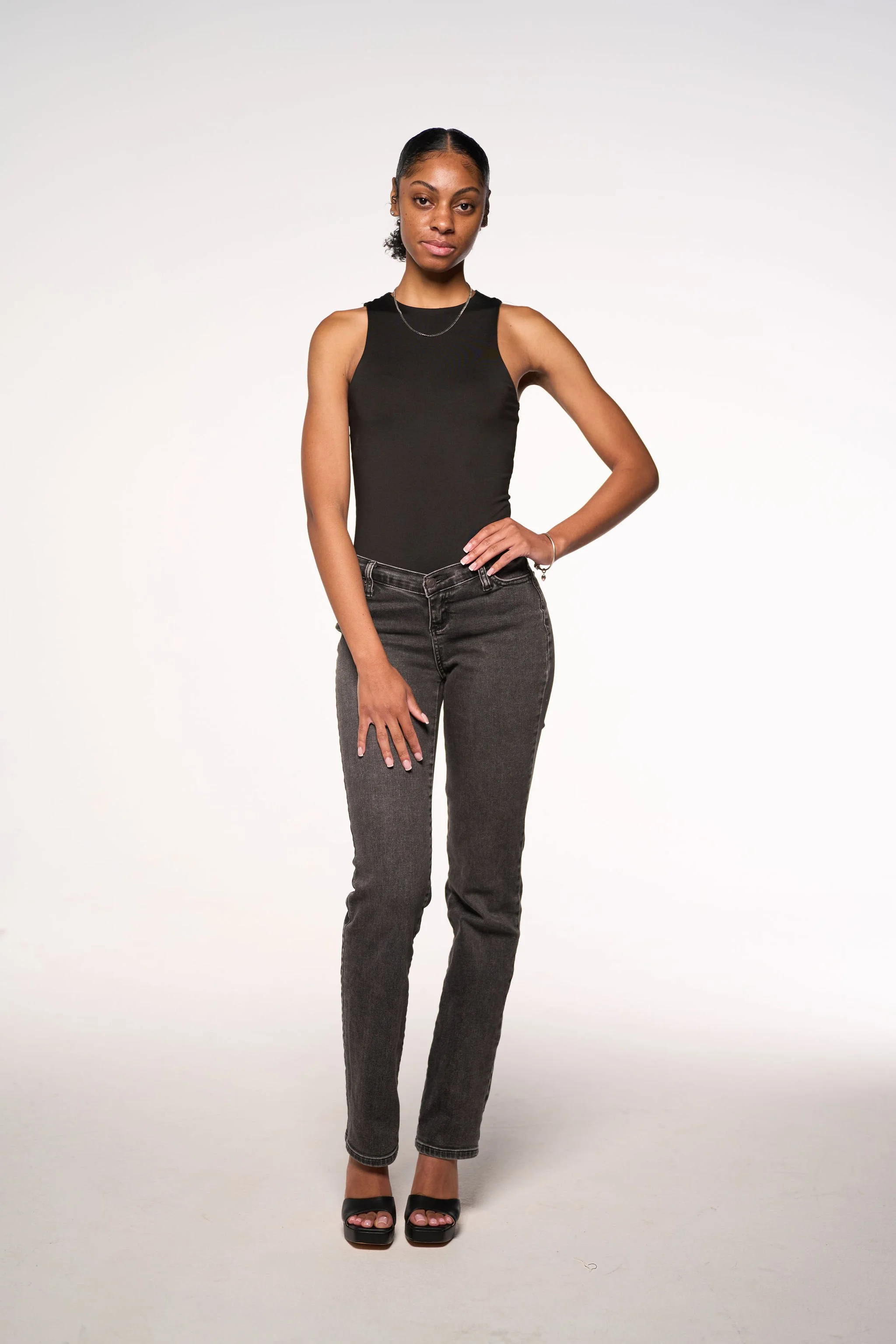 A young woman with dark hair slicked back, wearing a sleeveless black top, dark gray jeans, black open-toe heels, and minimal jewelry, standing against a plain white background with one hand on her hip.