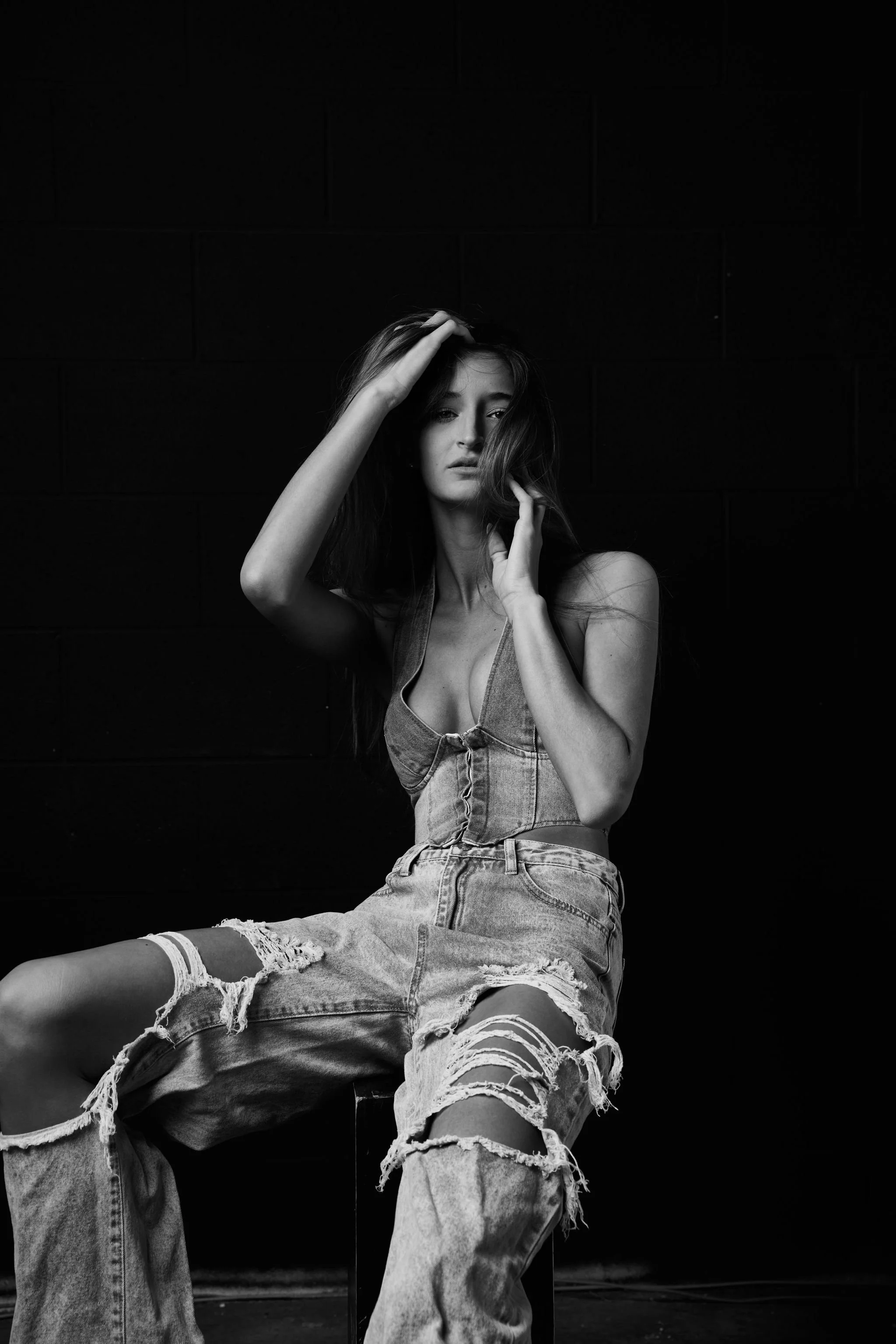 A black and white photo of a woman sitting against a dark wall, wearing ripped jeans and a denim top, touching her long hair and looking at the camera.