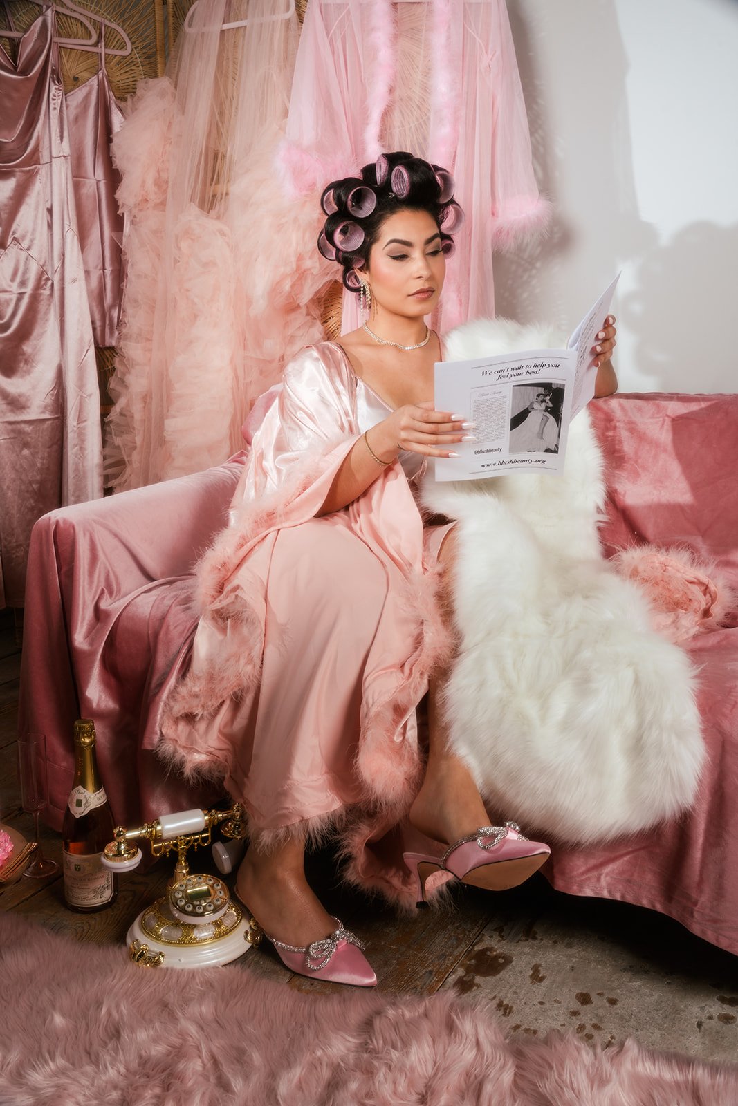 A woman in pink satin robes and curlers in her hair sits on a pink velvet couch, reading a magazine. She is surrounded by pink and white fluffy decor, including a couch blanket, dress, and a white faux fur pillow. There is a vintage rotary phone and a champagne bottle on the floor nearby.