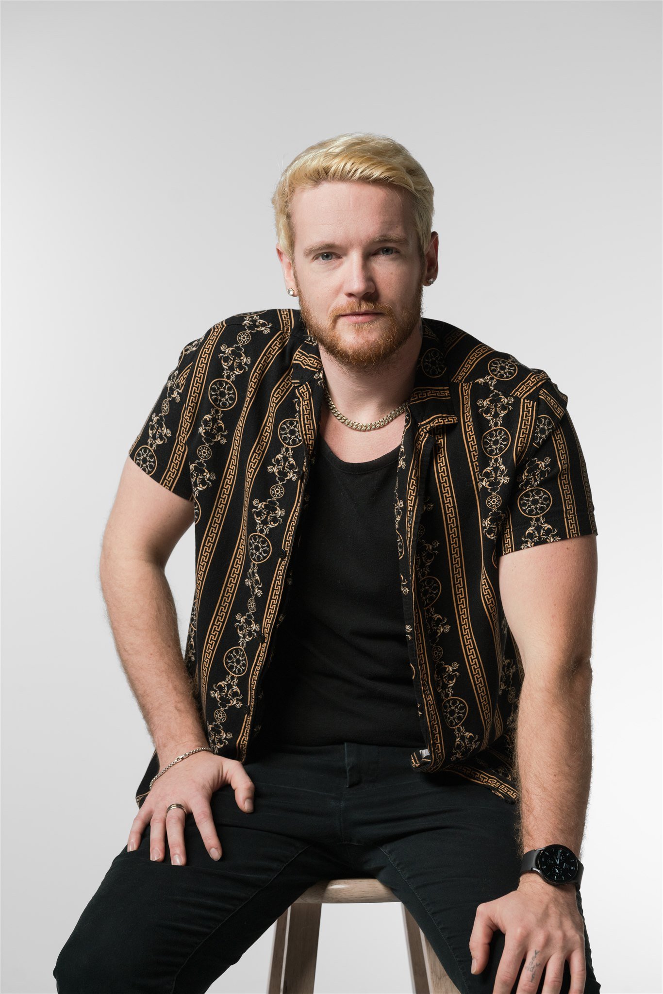 A young man with blonde hair, a beard, and earrings, wearing a black and gold patterned shirt over a black t-shirt, sitting on a wooden stool against a plain gray background.