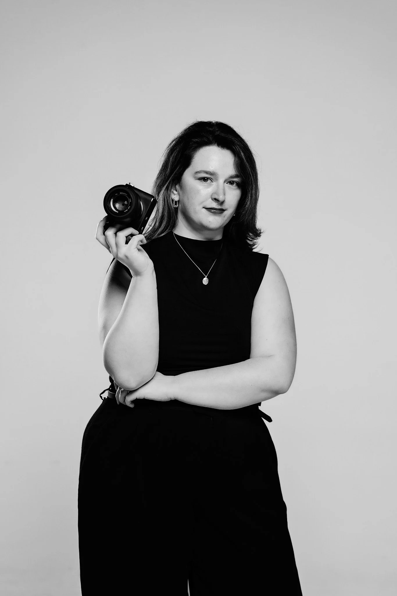 Black and white photo of a woman with shoulder-length hair, wearing a sleeveless top and necklace, holding a camera on her shoulder, against a plain background.