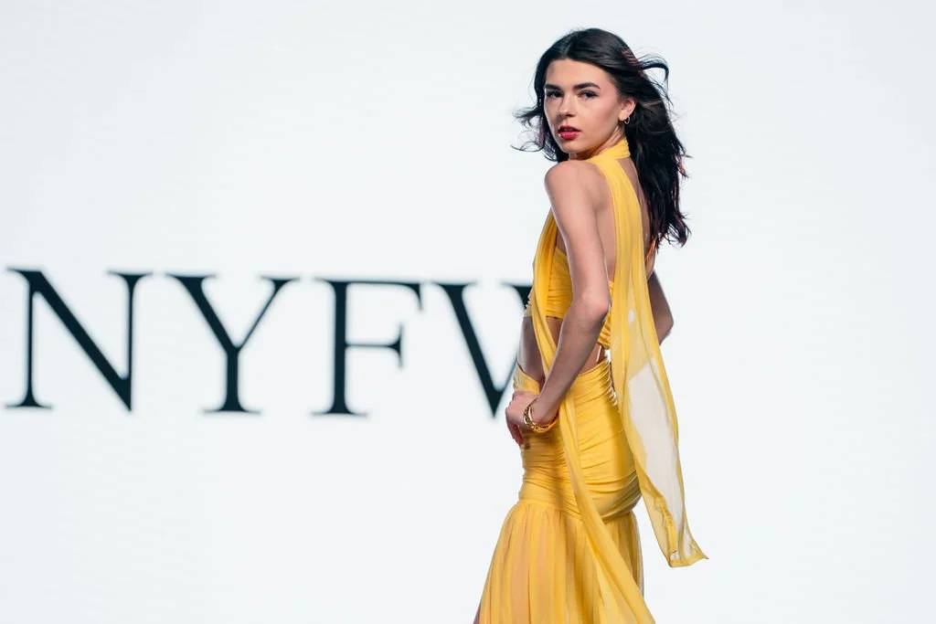 A woman in a yellow dress poses on a runway with the partial text 'NYFW' in the background.