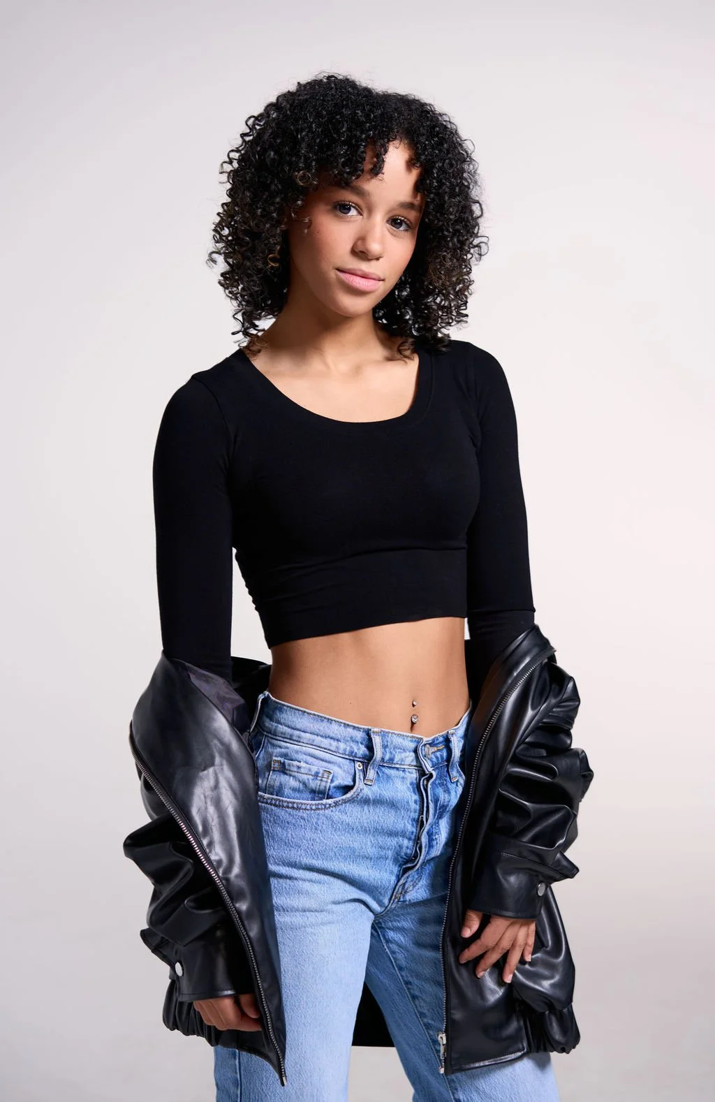 A young woman with black, curly hair wearing a black crop top, light blue jeans, and a black leather jacket draped off her shoulders, standing against a plain white background.