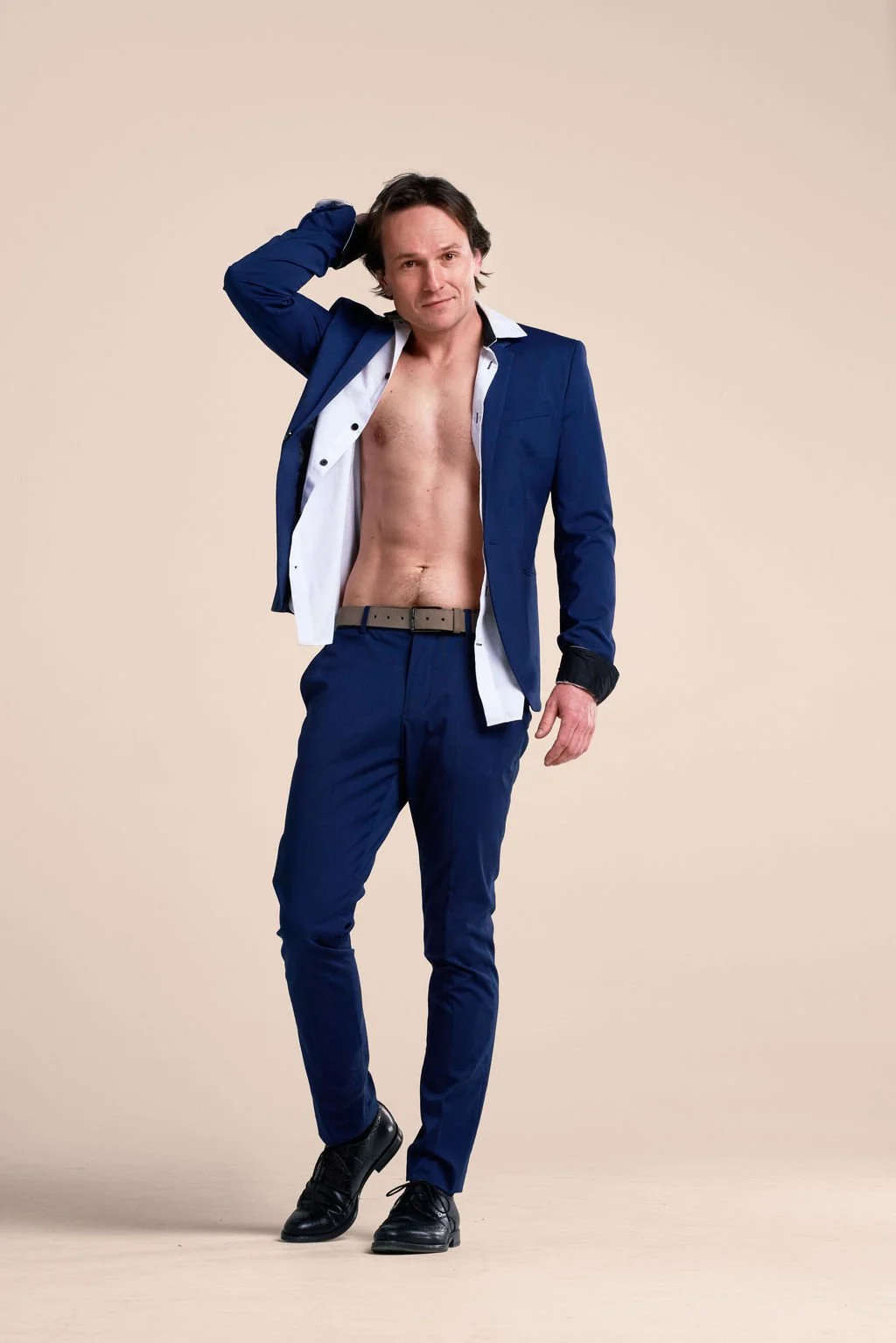 A man in a partially open blue suit, no shirt, standing with one hand on his head against a plain beige background.