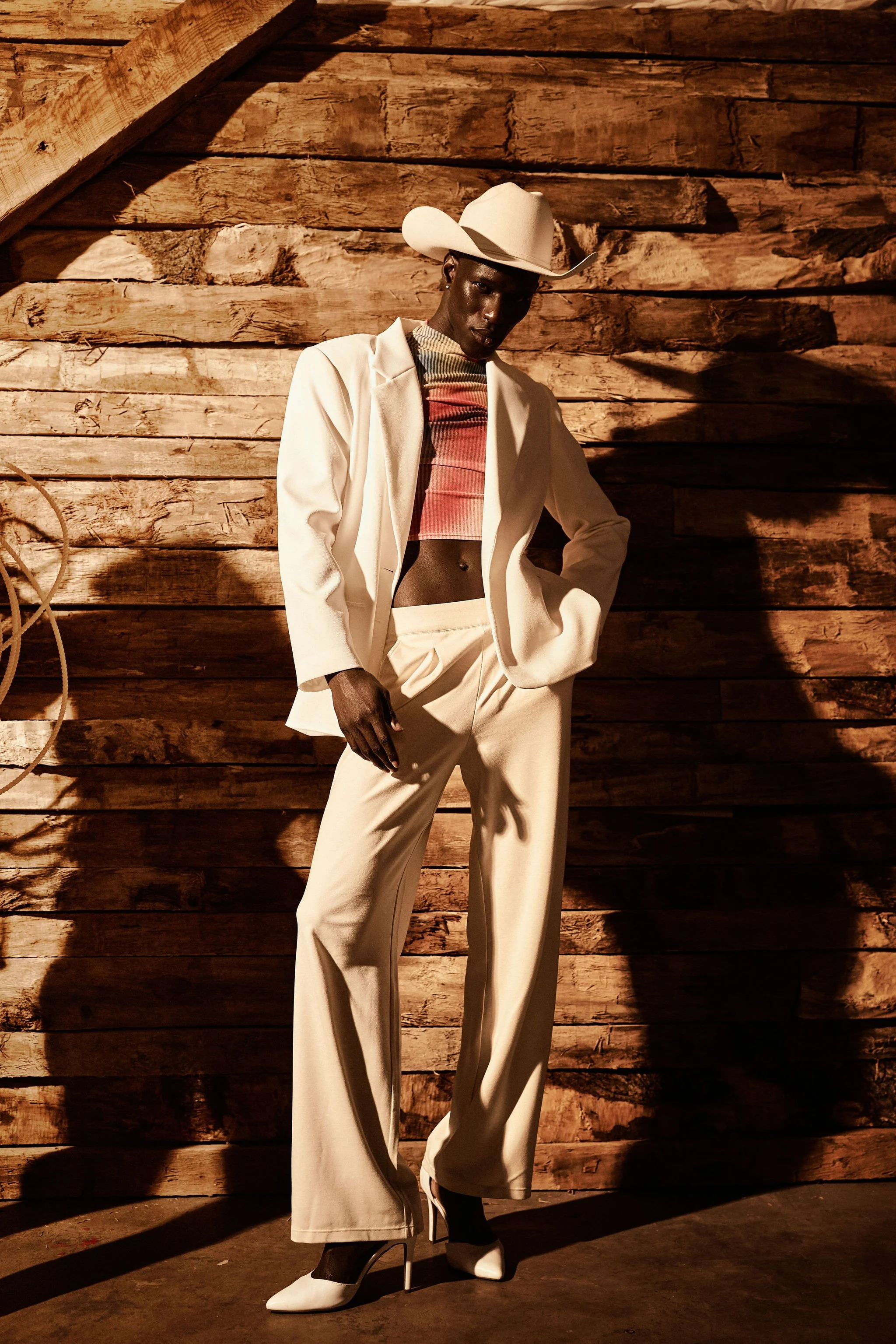 A person with dark skin in a white suit and cowboy hat standing against a wooden wall, wearing a colorful crop top and white high-heeled shoes.