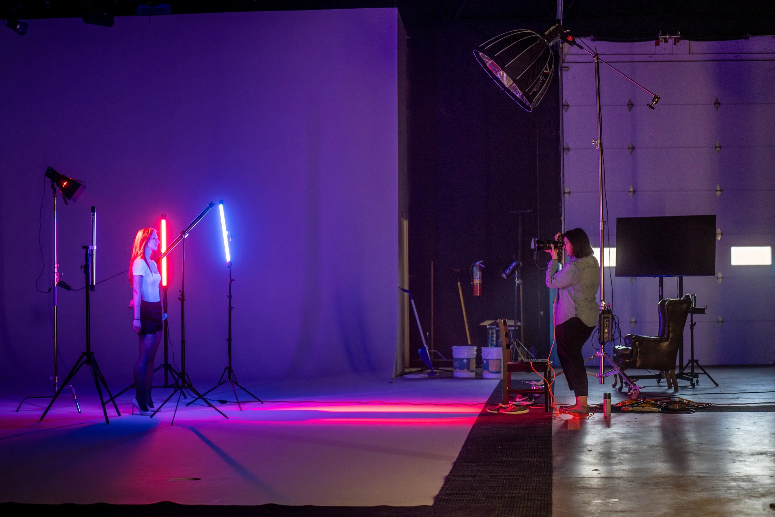 A woman standing in a photography studio with colorful neon lights, while a photographer takes her picture. The studio has a large backdrop, studio lighting, and equipment.