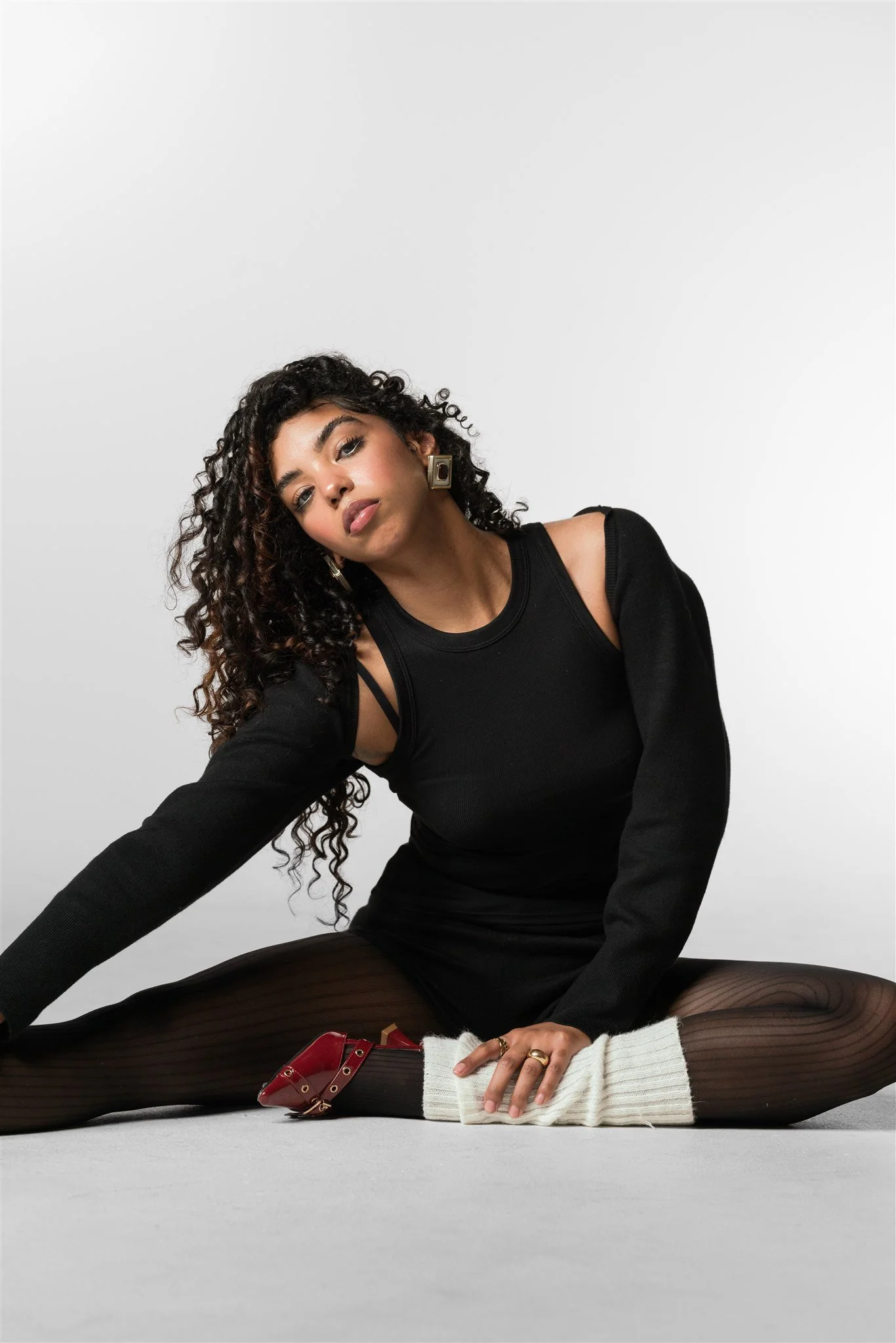 A woman with curly hair sitting on the floor against a plain white background, wearing a black top, black tights with a vertical stripe pattern, red shoes, a white knit sock, and gold earrings, with her head tilted slightly and looking at the camera.