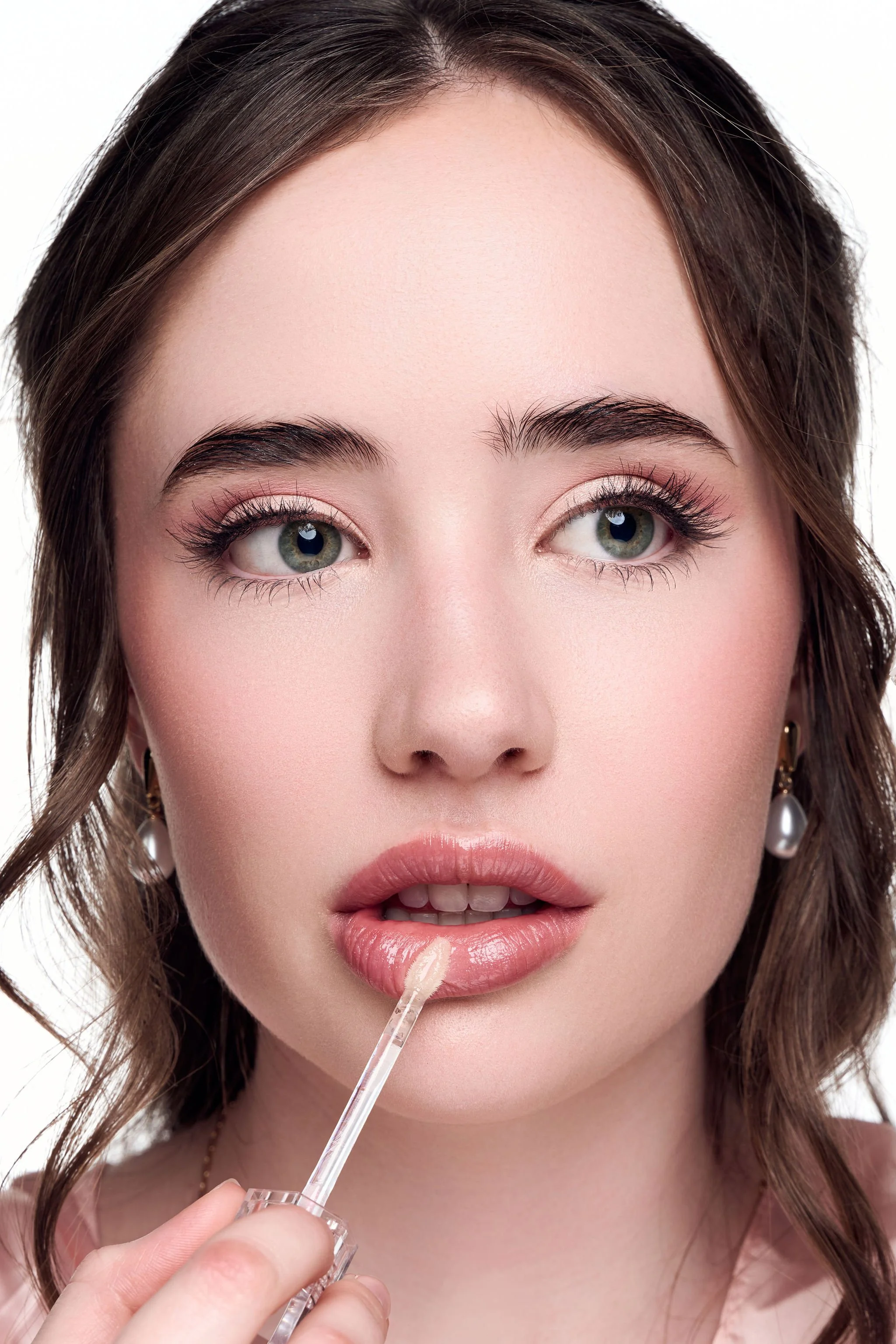 Close-up of a young woman with brown hair and green eyes, applying lip gloss with a brush while looking at the camera.