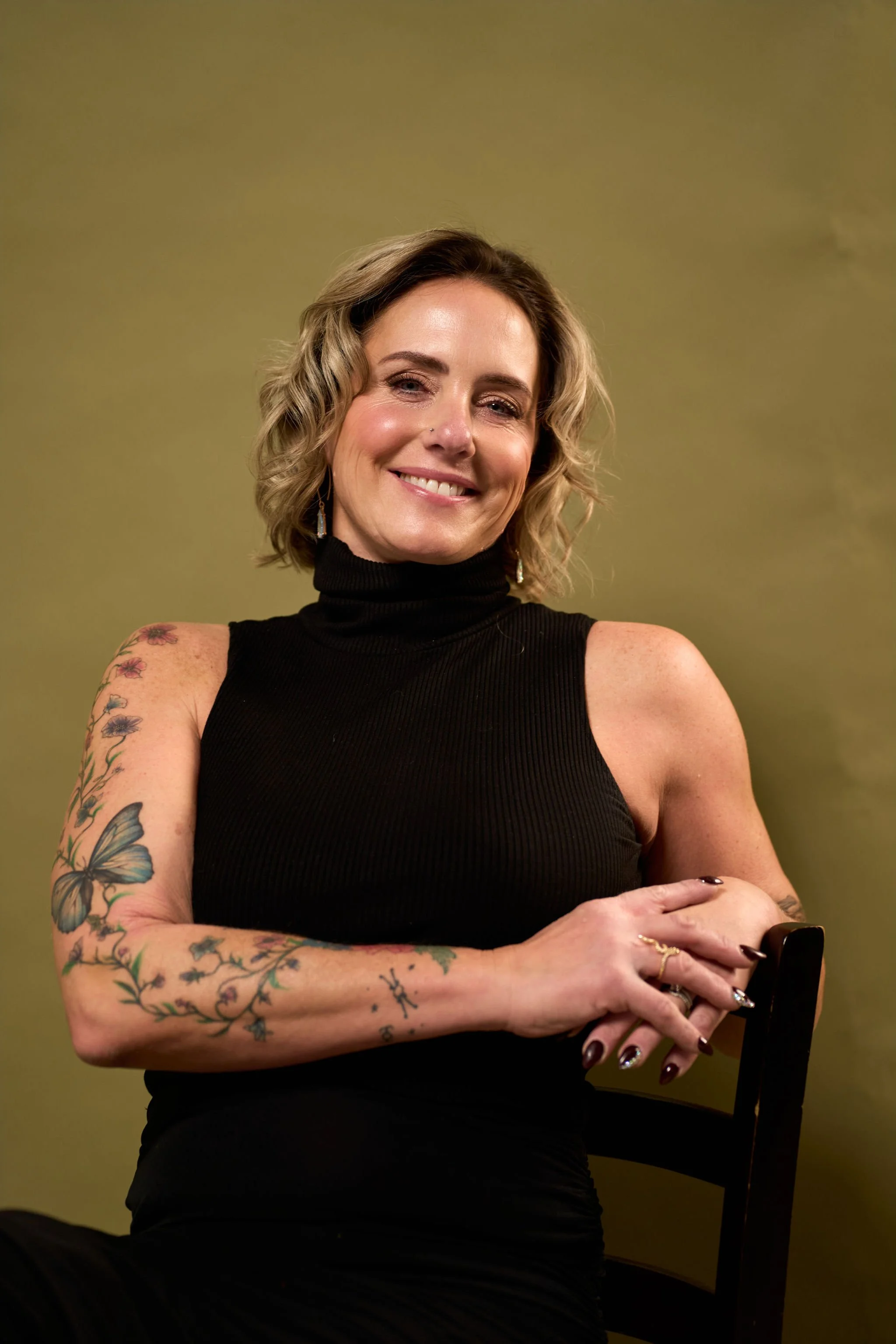 A smiling woman with short blonde hair, wearing a black sleeveless turtleneck, sitting on a chair against a muted green background. She has tattoos on her arms, dark nail polish, and earrings.