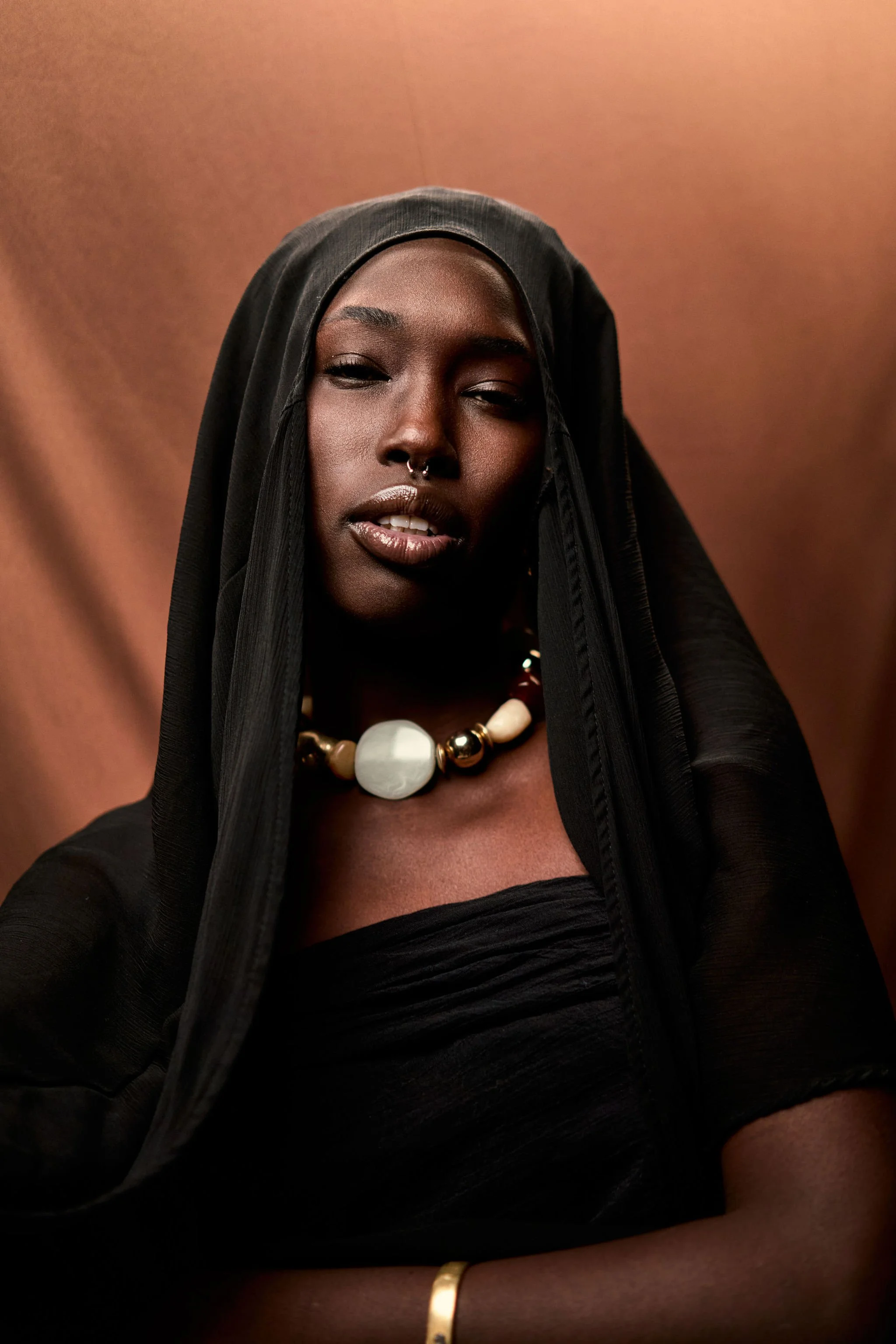 A woman with dark skin wearing a black headscarf, a black top, a chunky white and gold bead necklace, and a gold bracelet.