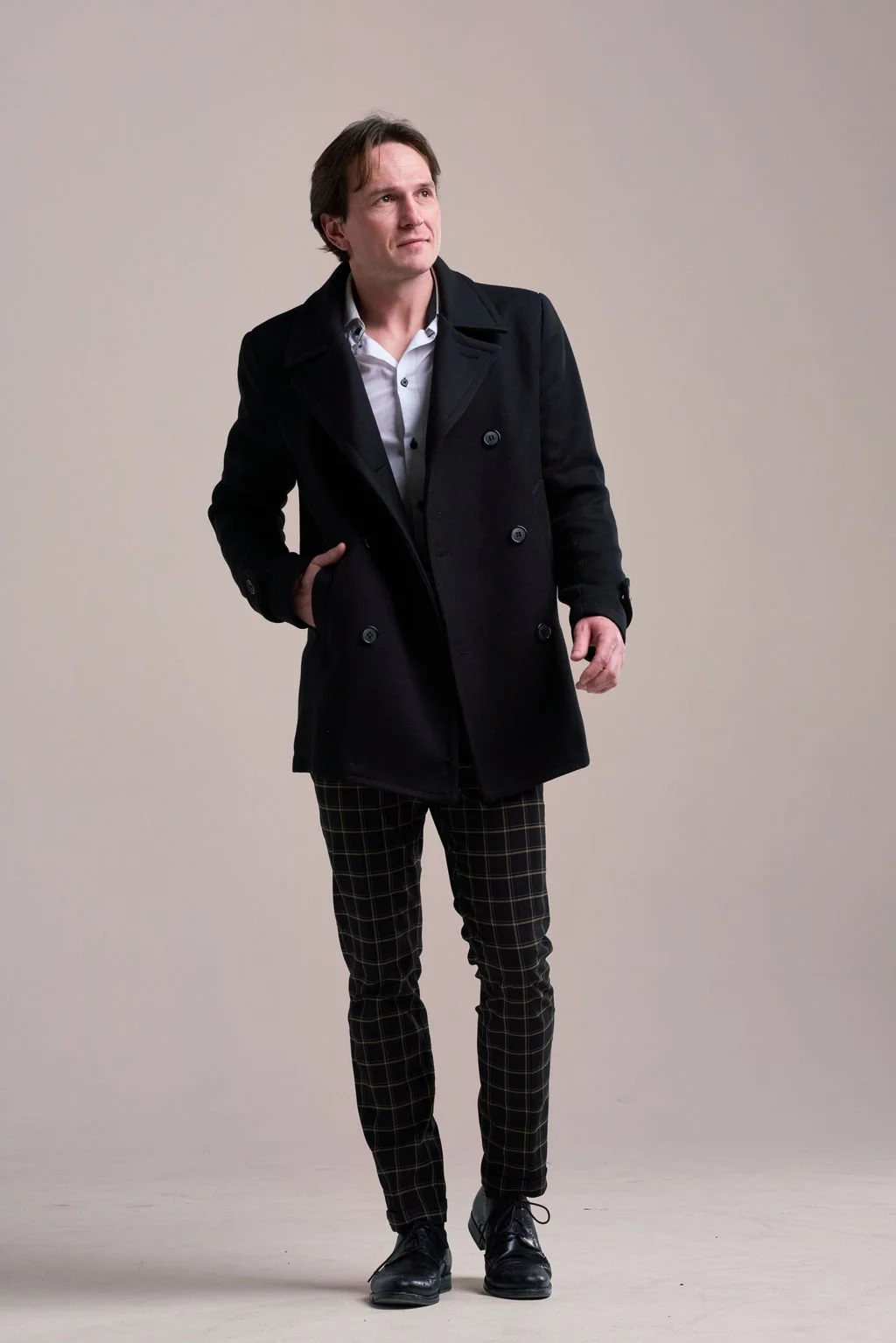 A man with brown hair wearing a black coat, white shirt, plaid pants, and black shoes, standing with one hand in his pocket against a plain beige background.