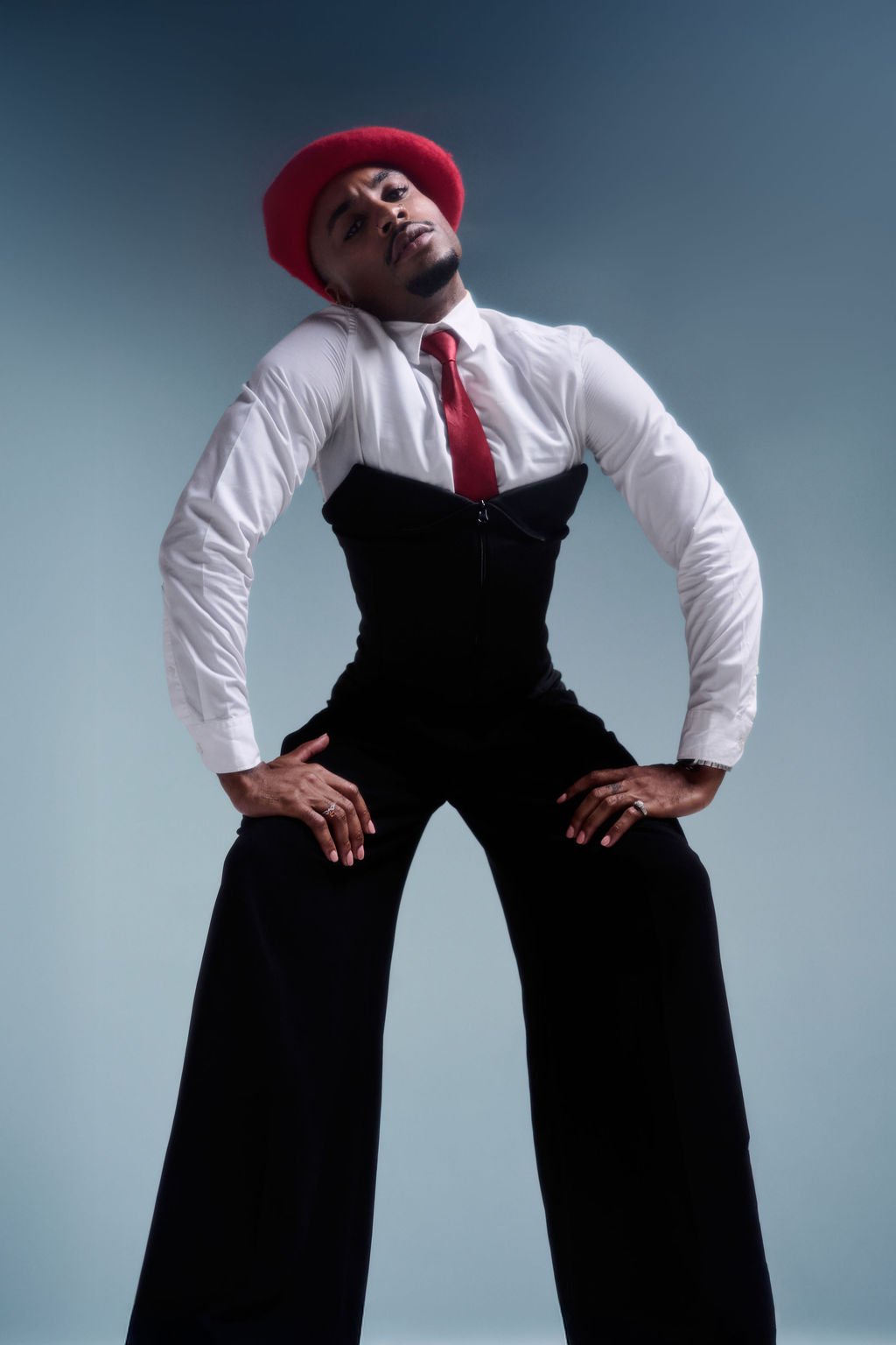 Person wearing a red hat, white shirt, red tie, black vest, and black wide-leg pants. The person is posing with hands on hips against a plain backdrop.