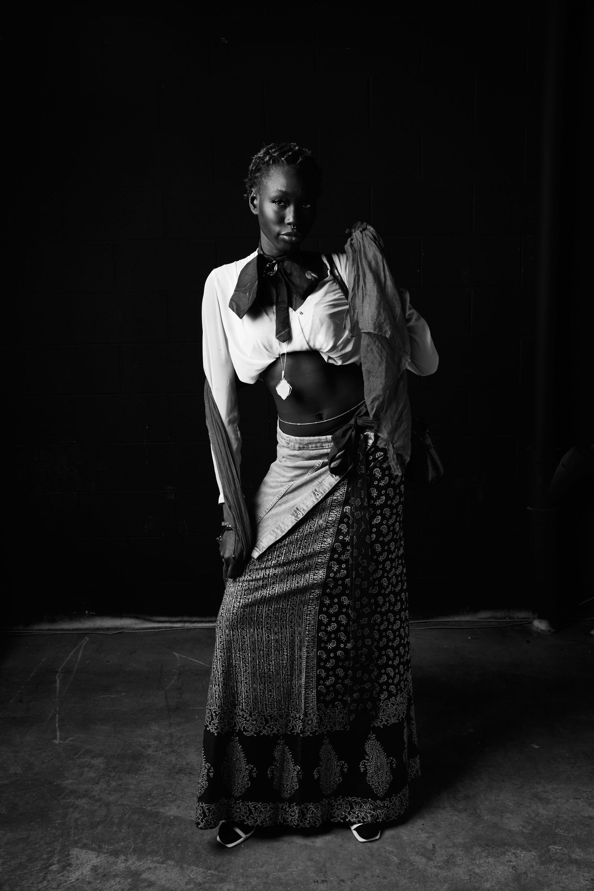 Black and white photo of a woman standing against a dark background, wearing a patterned skirt, a cropped top, and a shirt draped over her shoulder, with a confident pose.