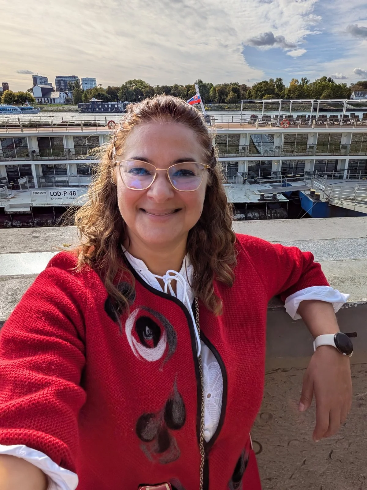 Maddy Caldaruse smiling in a red jacket on a river quay with a river cruise ship behind her