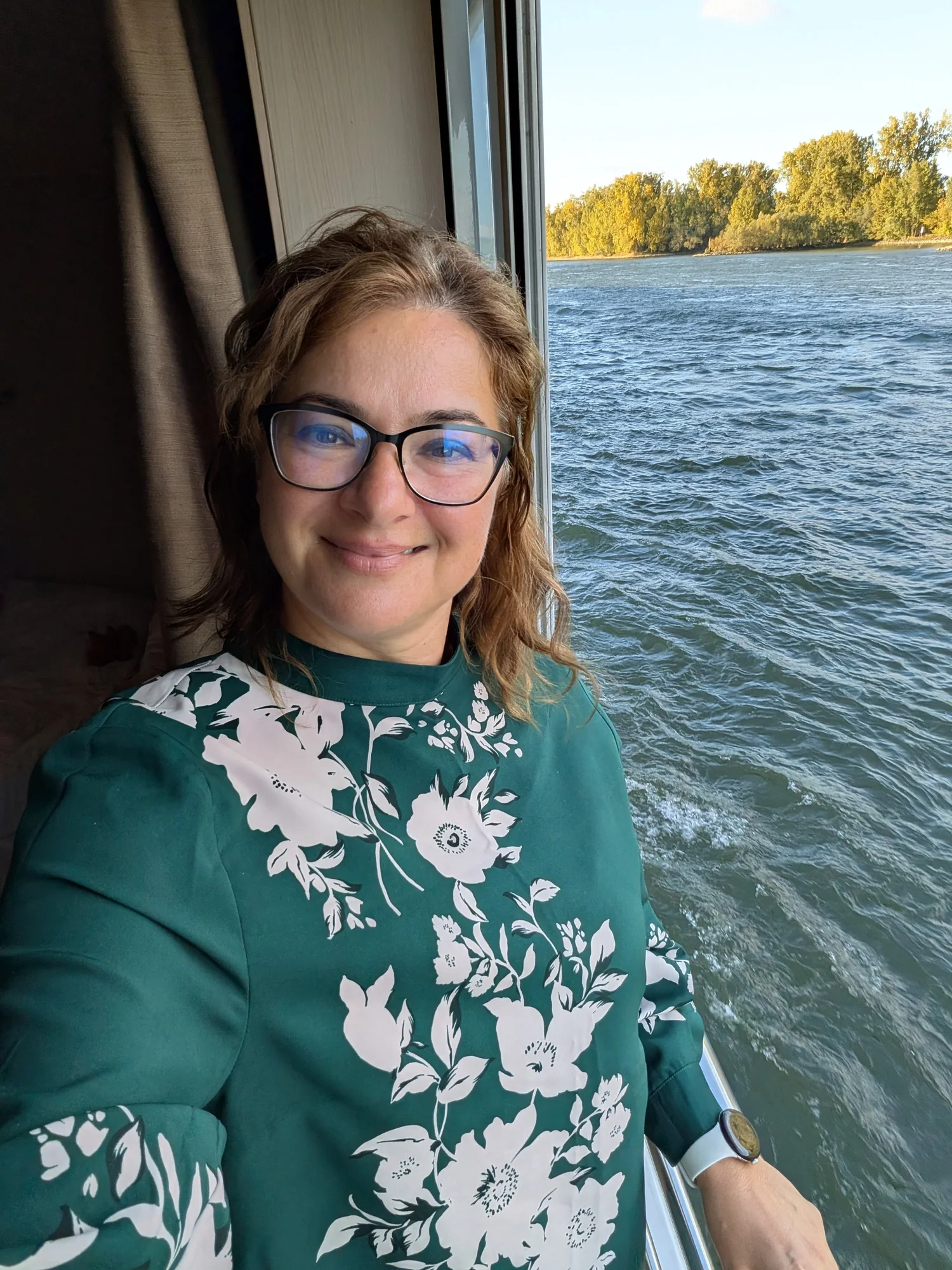 Maddy Căldărușe smiling by the window of a river cruise ship, with the river and autumn trees behind her