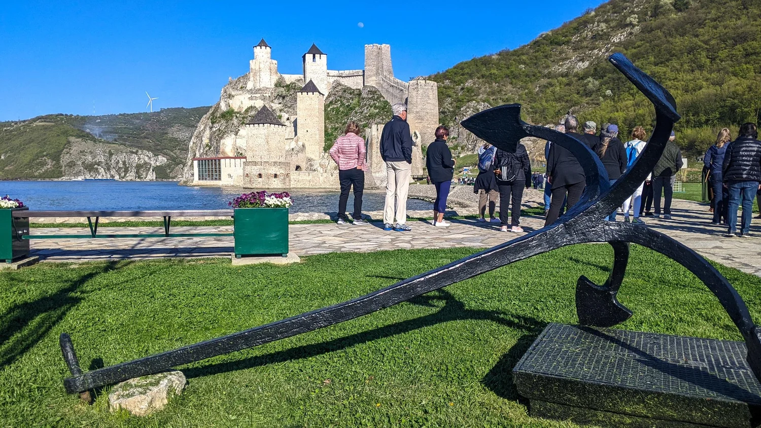 A group of travellers by a large anchor sculpture above the Danube, with Golubac Fortress behind them