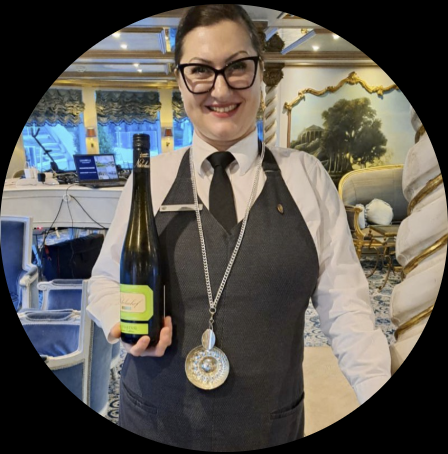 Mila smiling and holding a bottle of wine in an elegant, decorated room with blue lighting and ornate furniture.