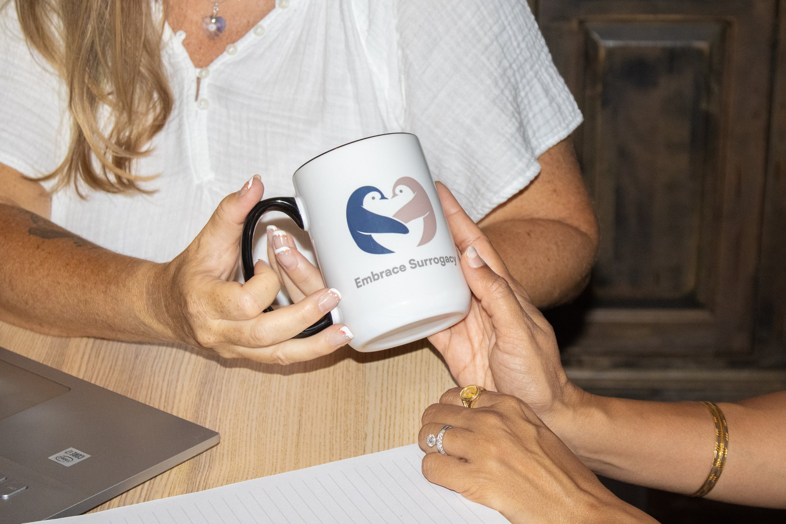 Two women exchanging a white mug with a logo of two swans and the text 'Embrace Surrogacy' in an indoor setting, with one woman wearing a white shirt and the other with jewelry on her hands.