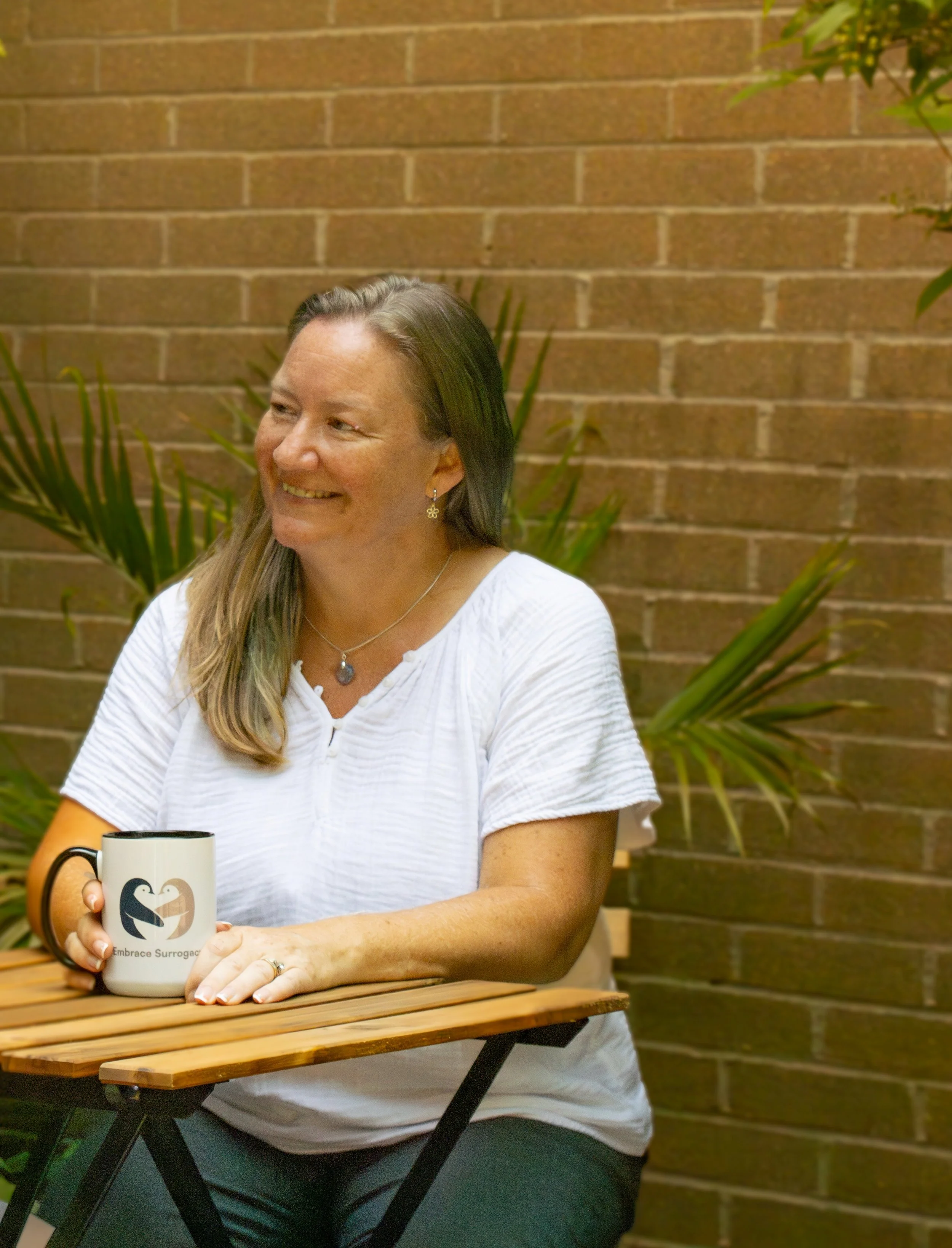 A woman with long blonde hair smiling and sitting at a wooden table, holding a mug with a black and white logo, in front of a brick wall with green plants.