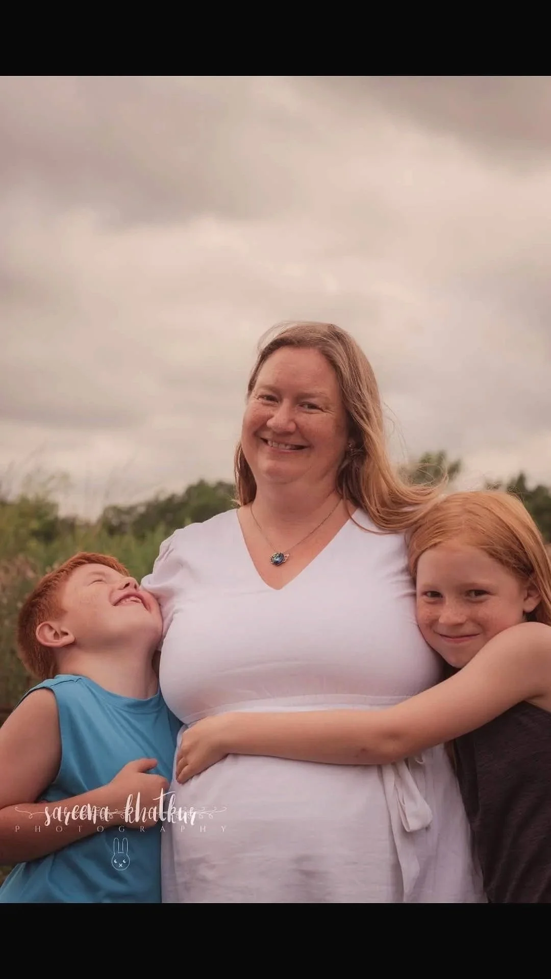 A smiling woman with two children hugging her outdoors on a cloudy day.