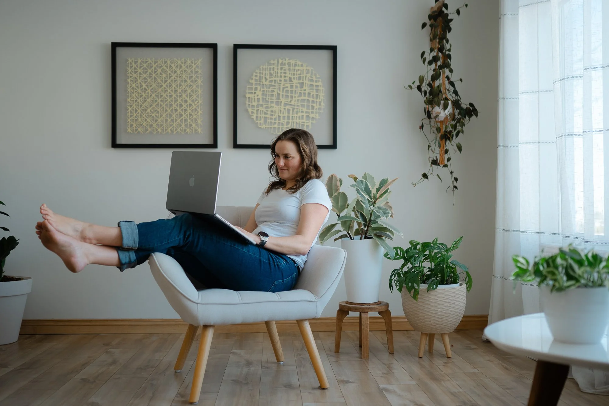 Une femme assise confortablement dans un fauteuil, utilisant un ordinateur portable sur ses jambes, entourée de nombreuses plantes d'intérieur, dans une pièce lumineuse avec des œuvres d'art abstraites aux murs.