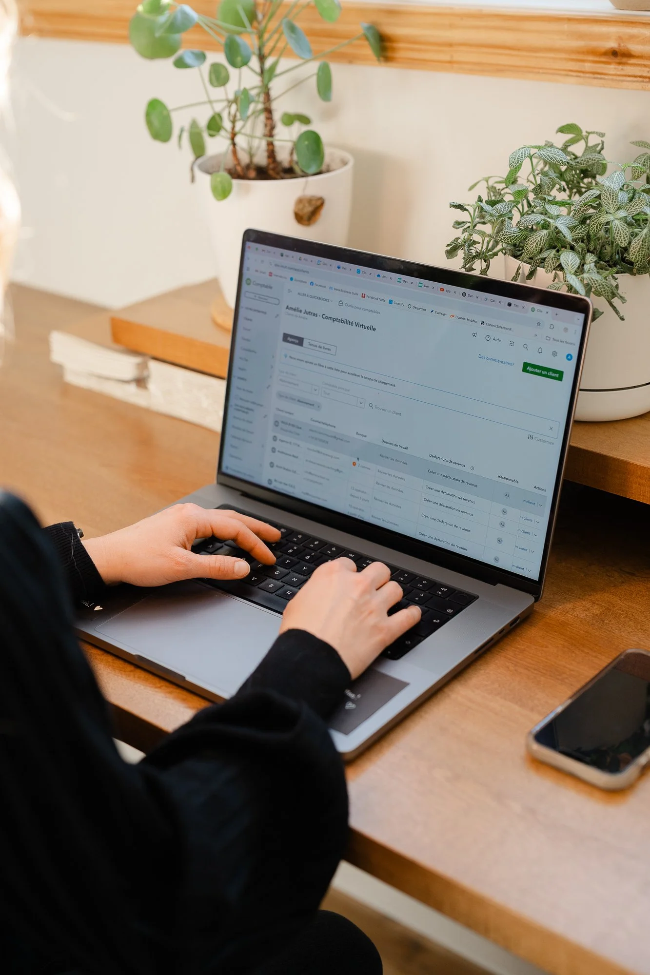 Une personne utilisant un ordinateur portable à un bureau en bois, avec des plantes en pot en arrière-plan.