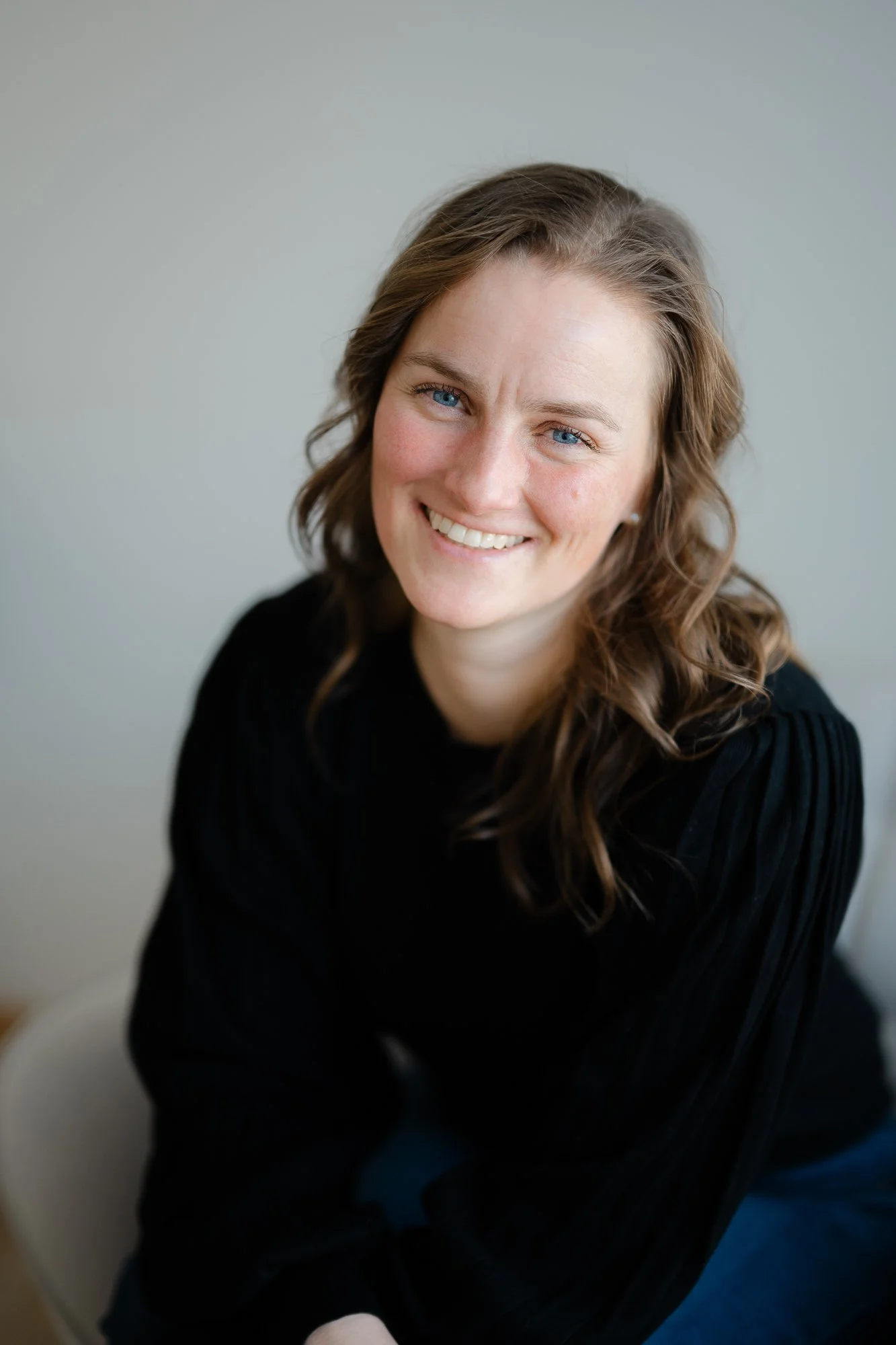 Une femme souriante avec des cheveux bruns ondulés, portant un pull noir, assise dans une pièce avec un mur clair en arrière-plan.