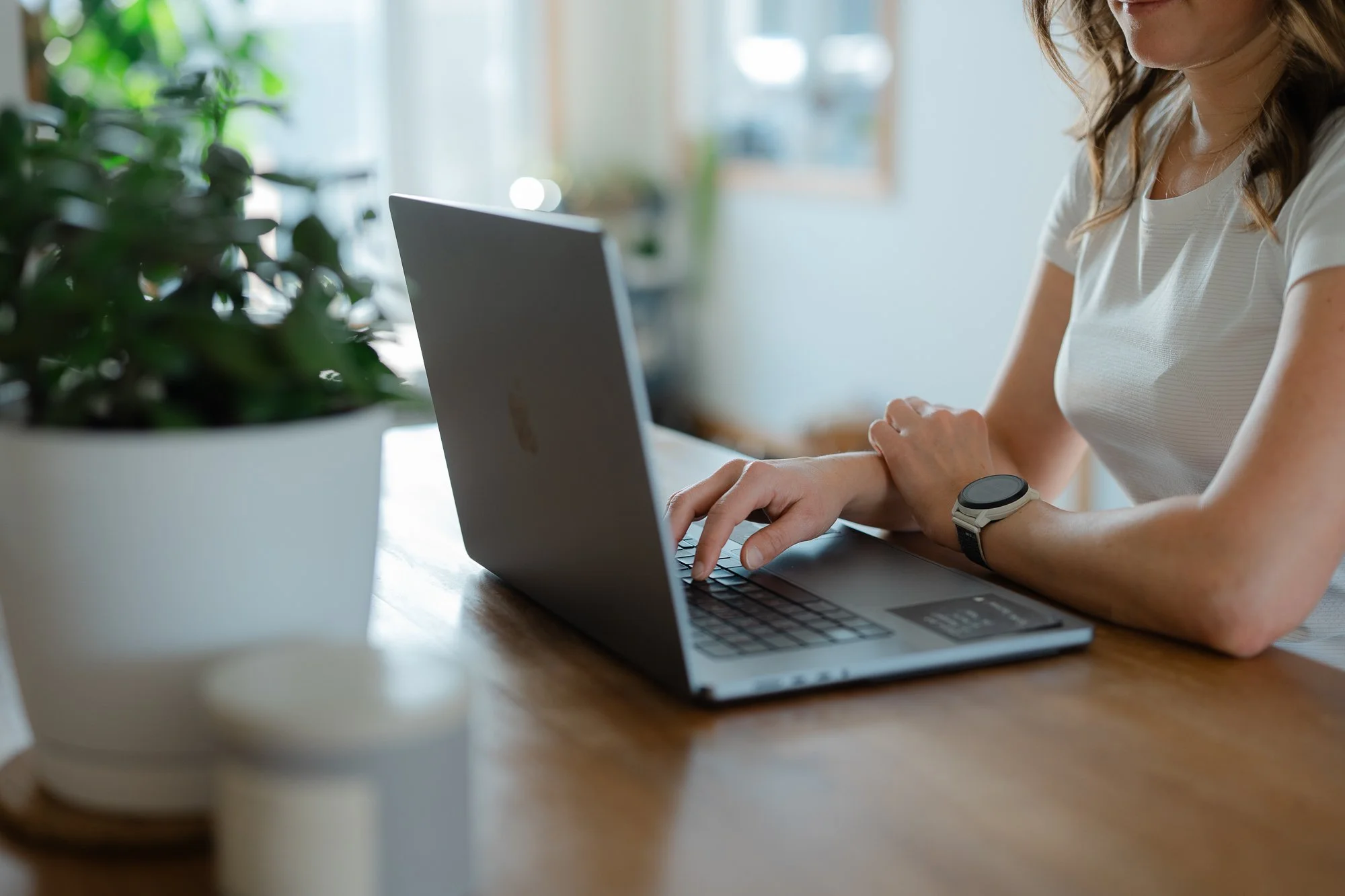 Une femme travaille sur un ordinateur portable sur une table en bois, avec une plante verte floue en premier plan et une pièce lumineuse en arrière-plan.