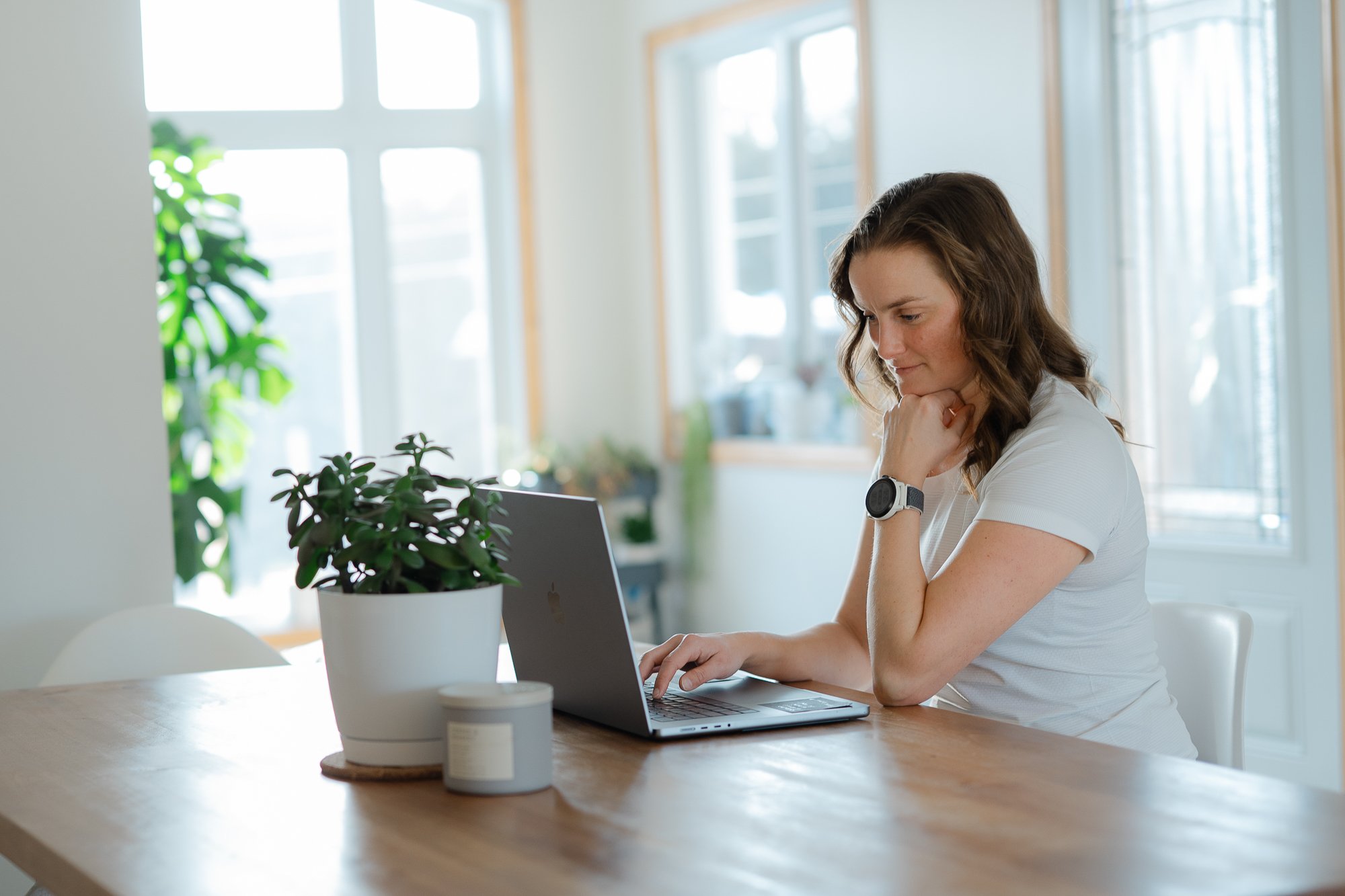Une femme aux cheveux bruns courts, portant un t-shirt blanc et une montre, travaille sur un ordinateur portable sur une table en bois clair dans une pièce lumineuse avec de grandes fenêtres et des plantes d'intérieur.