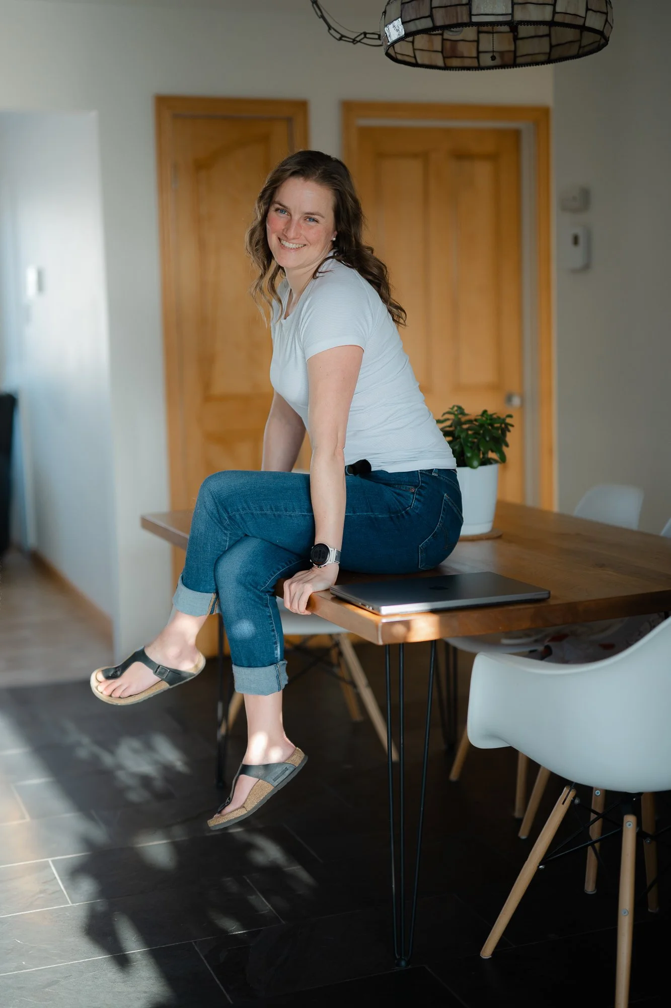 Une femme souriante au salon, assise sur une table en bois, portant un t-shirt blanc, jeans, sandales, avec une plante en pot derrière elle, dans une pièce lumineuse.