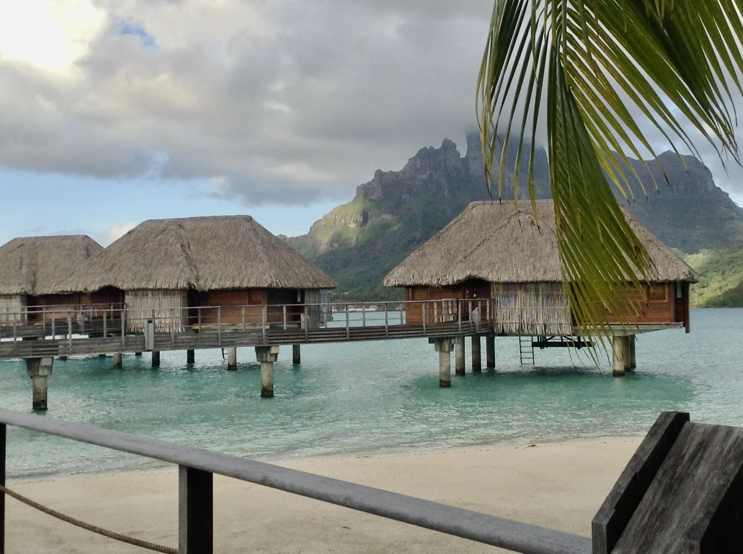 Overwater bungalows with thatched roofs over turquoise water, backed by lush mountains and partly cloudy sky, with a palm tree in the foreground.
