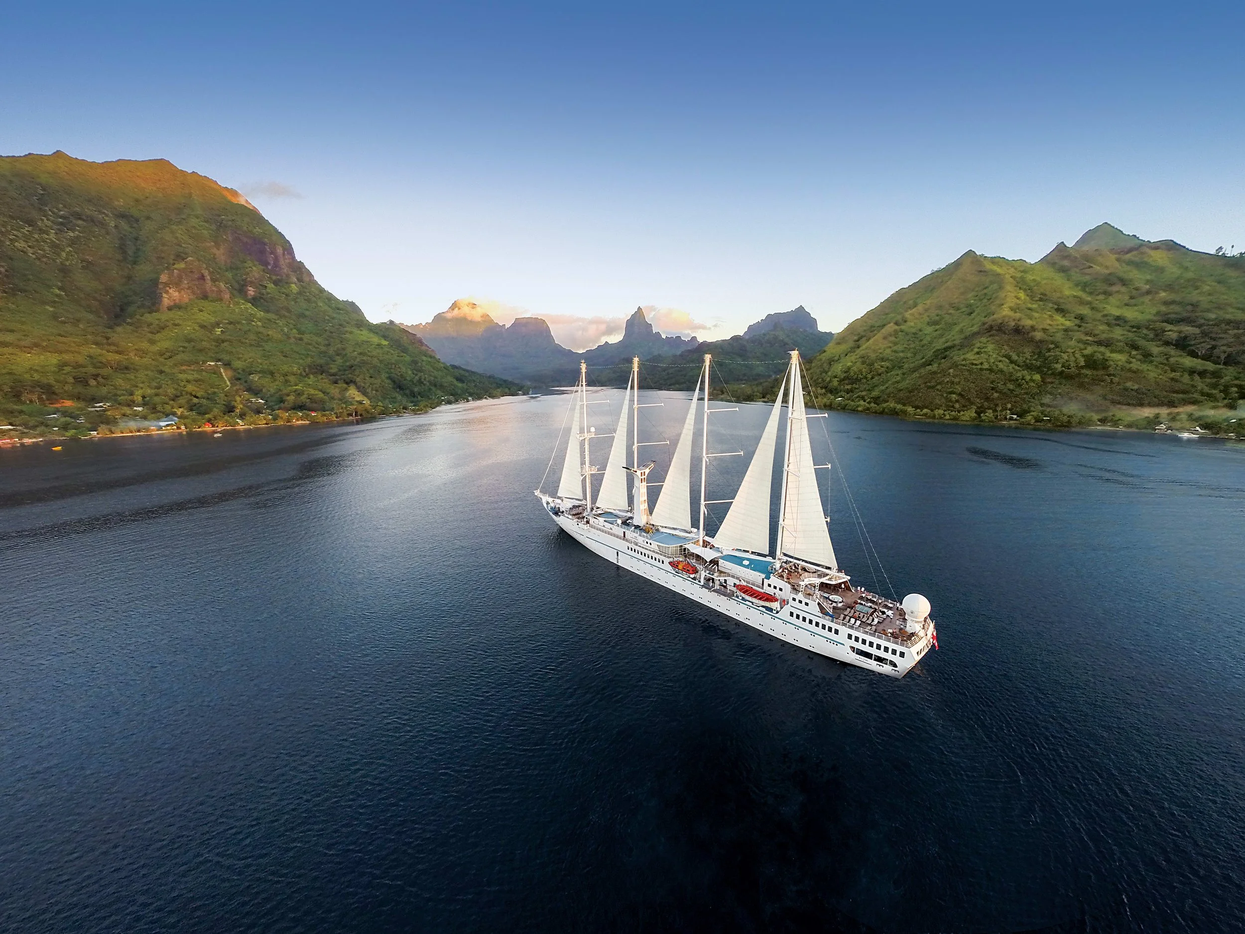 A white sailing cruise ship with multiple sails on a river, surrounded by green mountains under a clear blue sky.