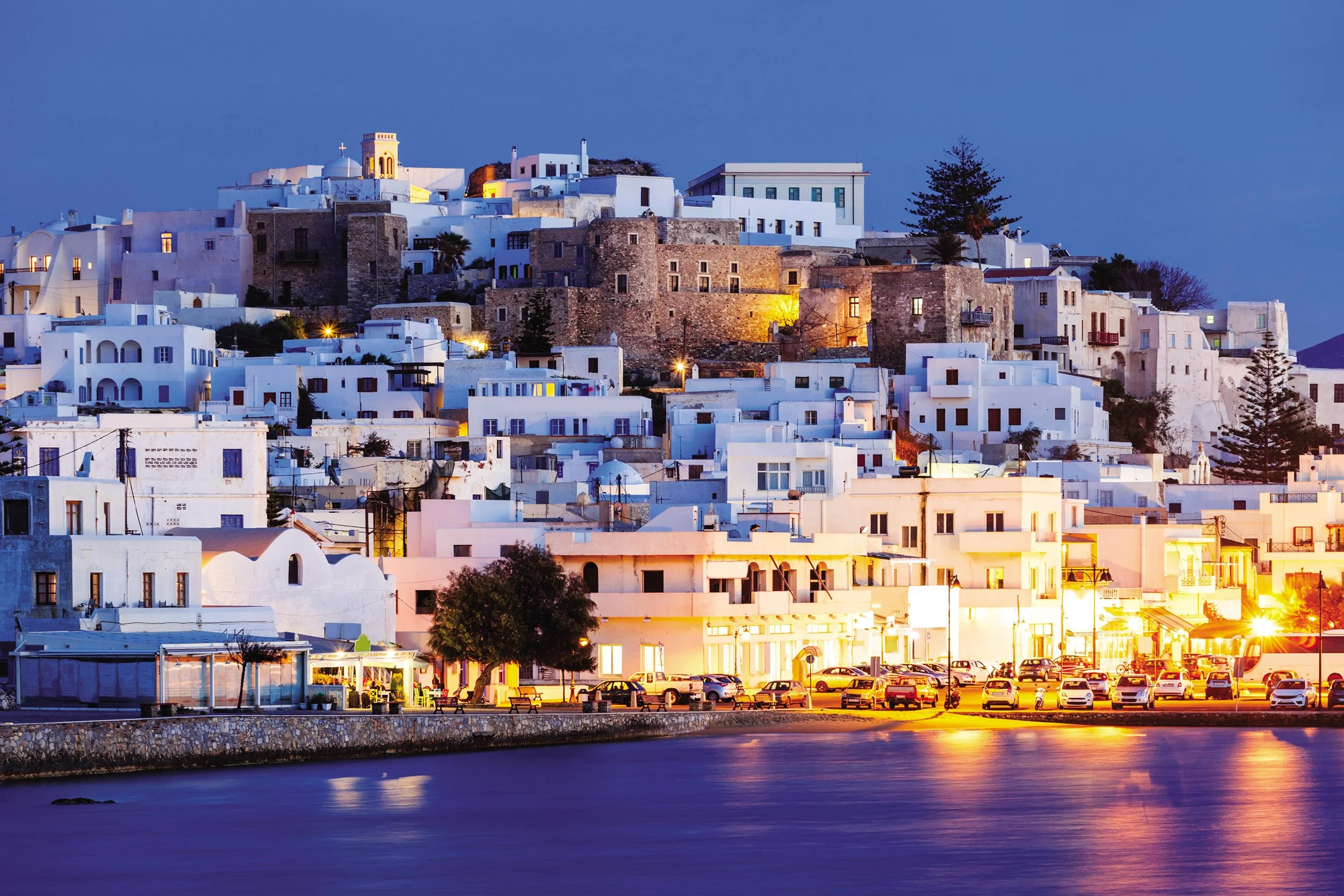 A view of a hillside town at dusk with white buildings, some with blue accents, and a waterfront with parked cars and water reflecting lights.