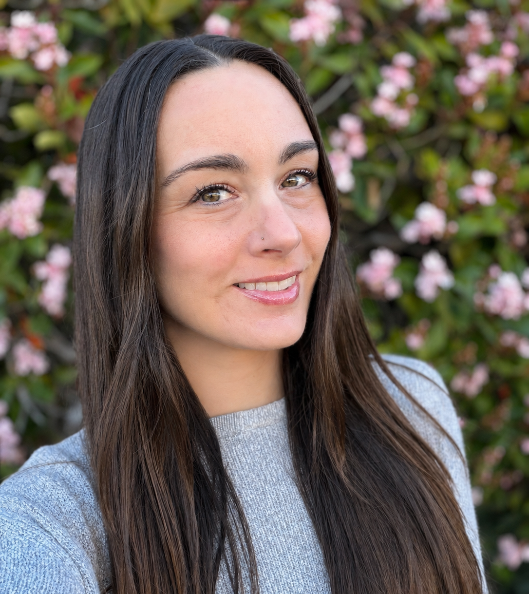 A woman with long dark hair smiling outdoors, wearing a black top and a silver necklace, with green plants and a tree in the background.