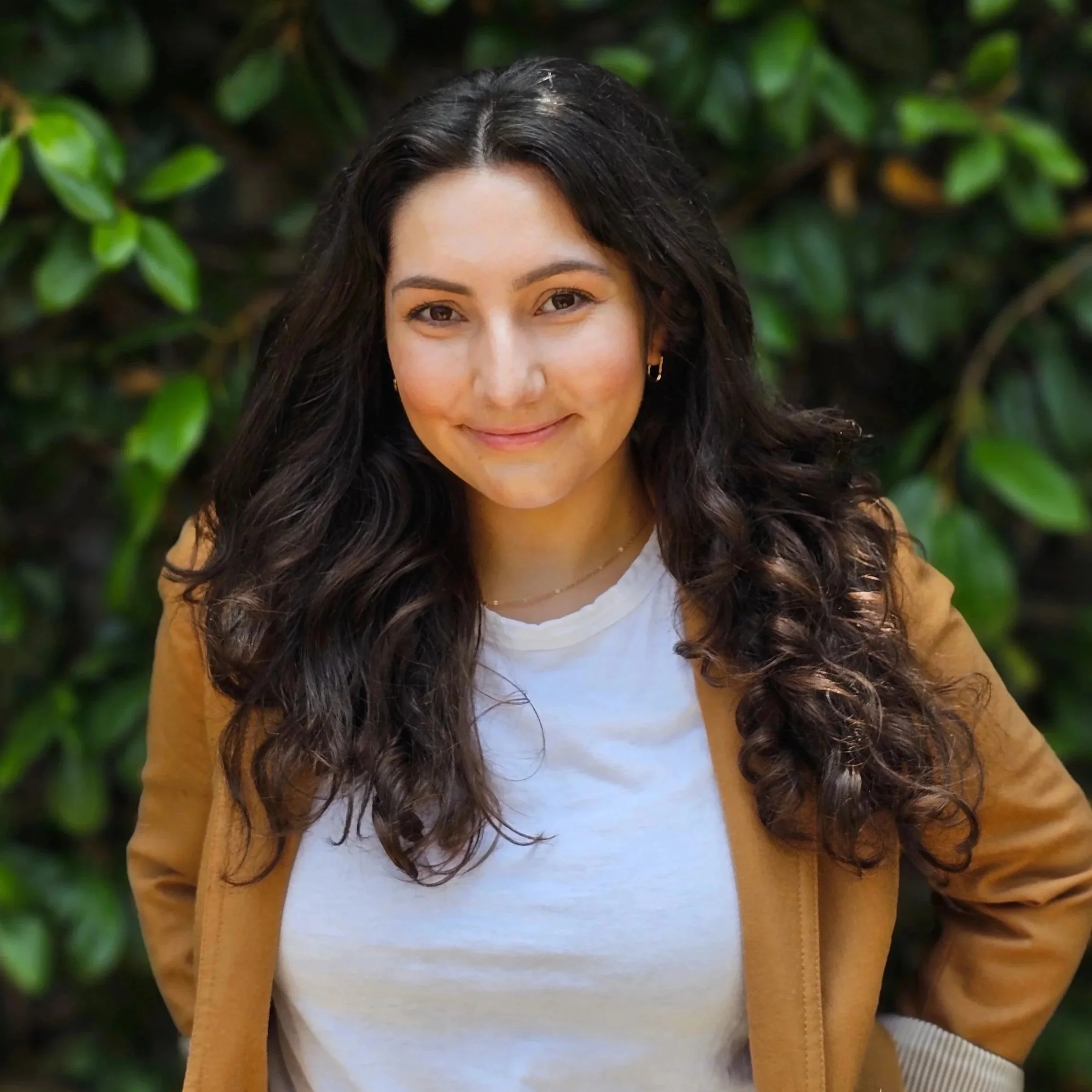 Young woman with dark curly hair smiling outdoors with green leaves in the background.