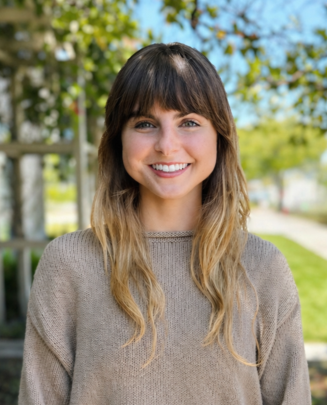 A young woman with long wavy brown hair and bangs is smiling, wearing a light pink top and layered necklaces, standing in front of a plain wall with two windows letting in natural light.