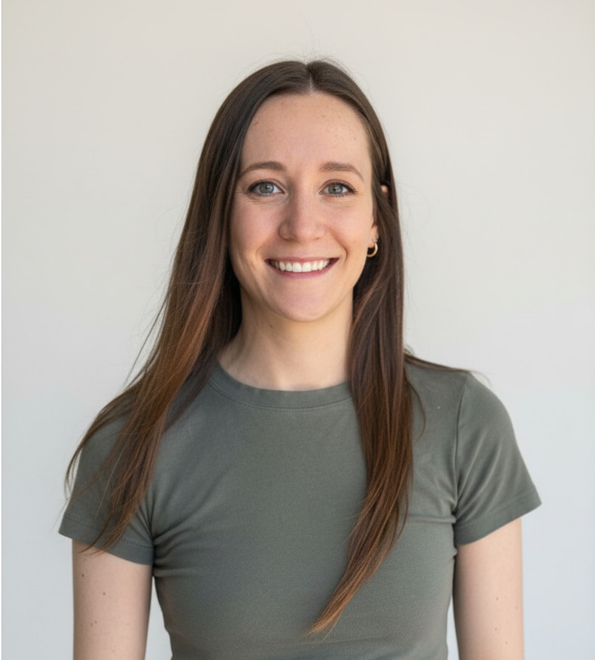Young woman with long brown hair, wearing a black top, smiling, standing against a plain white background.