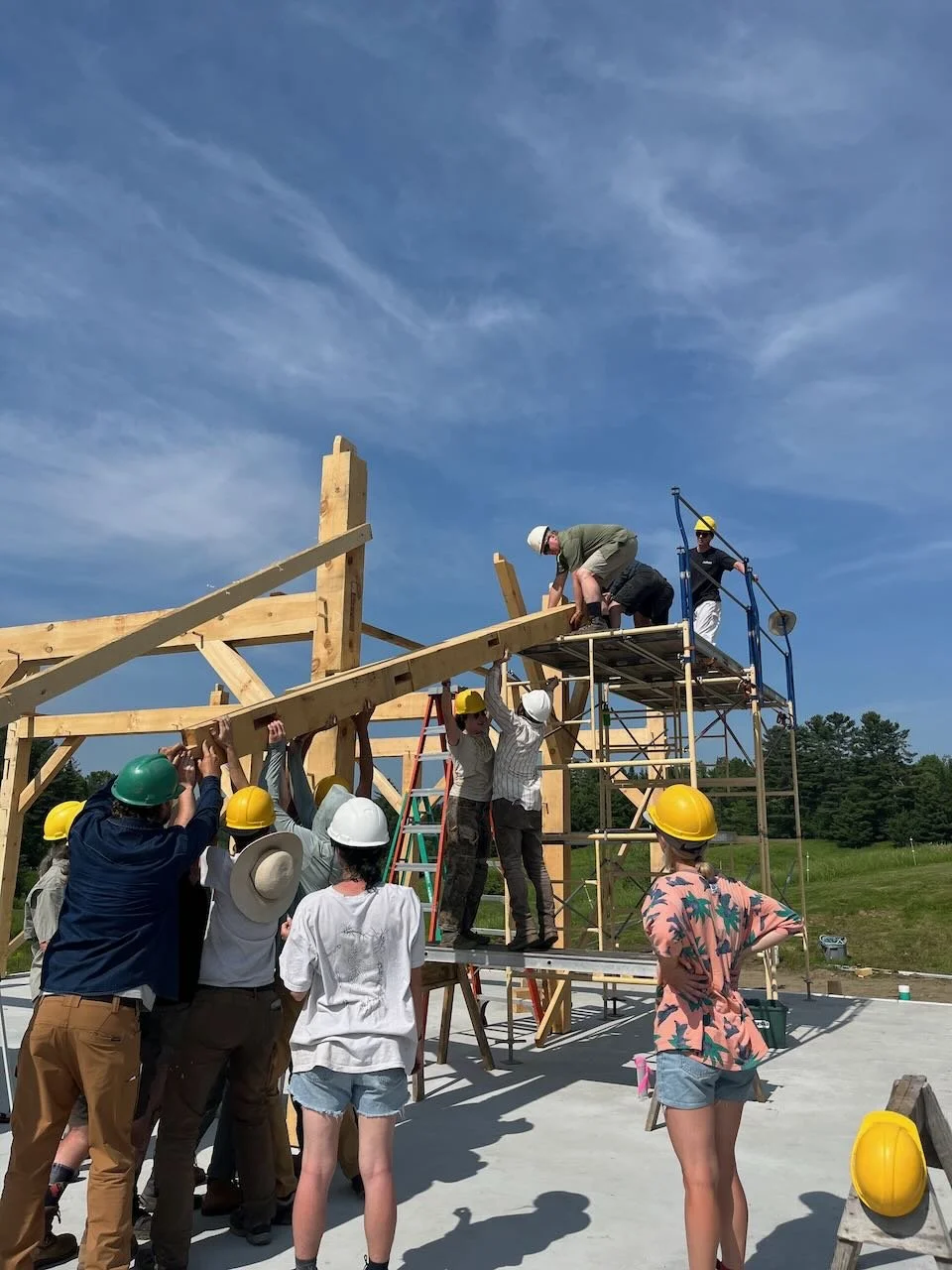 Timber framing students pass a heavy beam up to a team standing on scaffolding