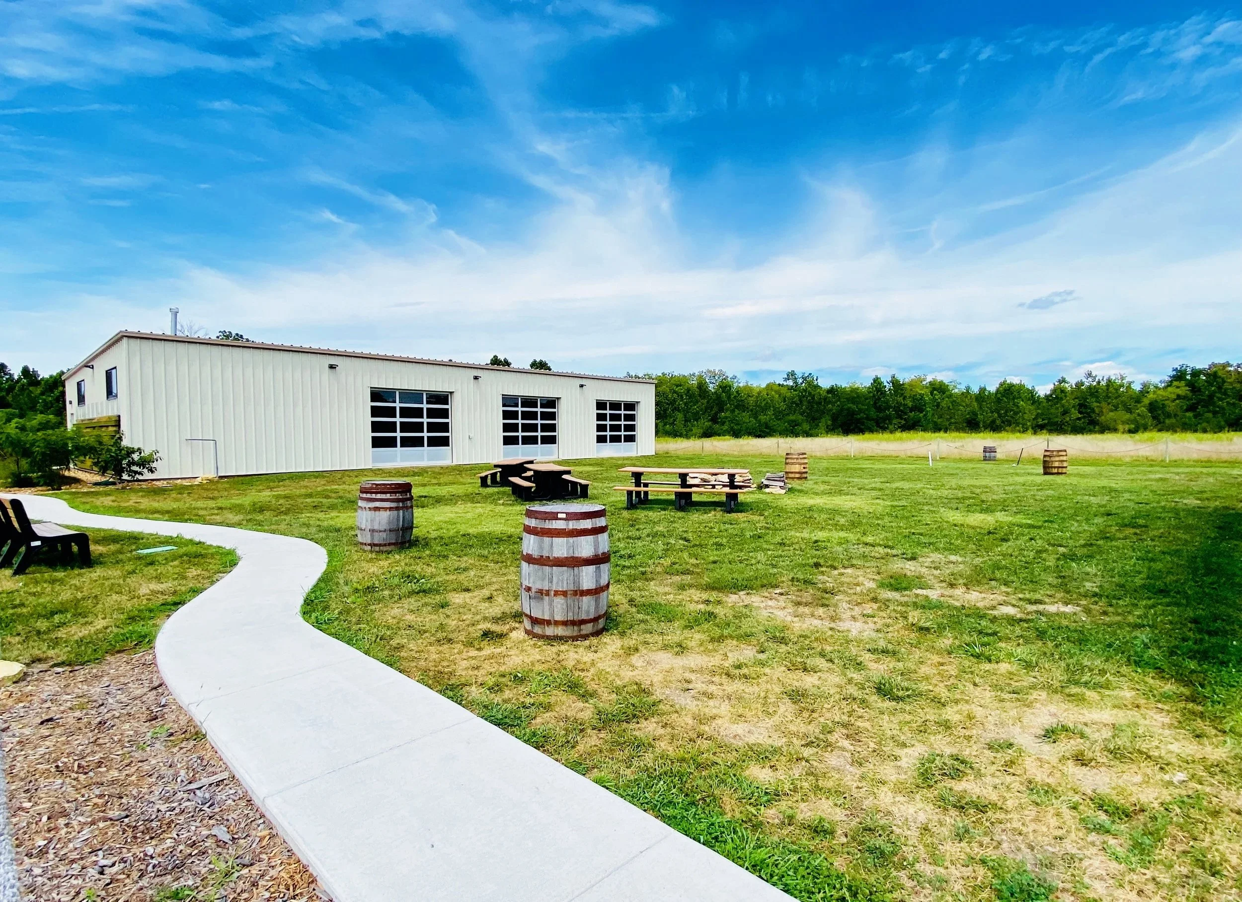 A large white building in a grassy outdoor area with picnic tables and barrels, under a partly cloudy blue sky.