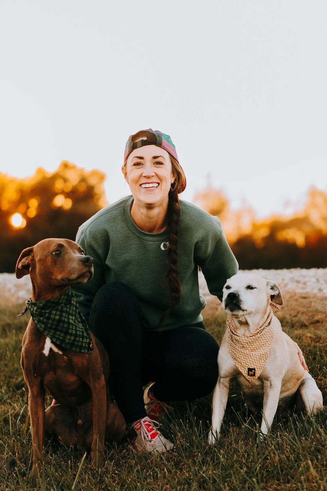 A woman smiling and crouching on the grass outdoors in the evening, accompanied by two dogs, one brown and one white, both wearing bandanas.