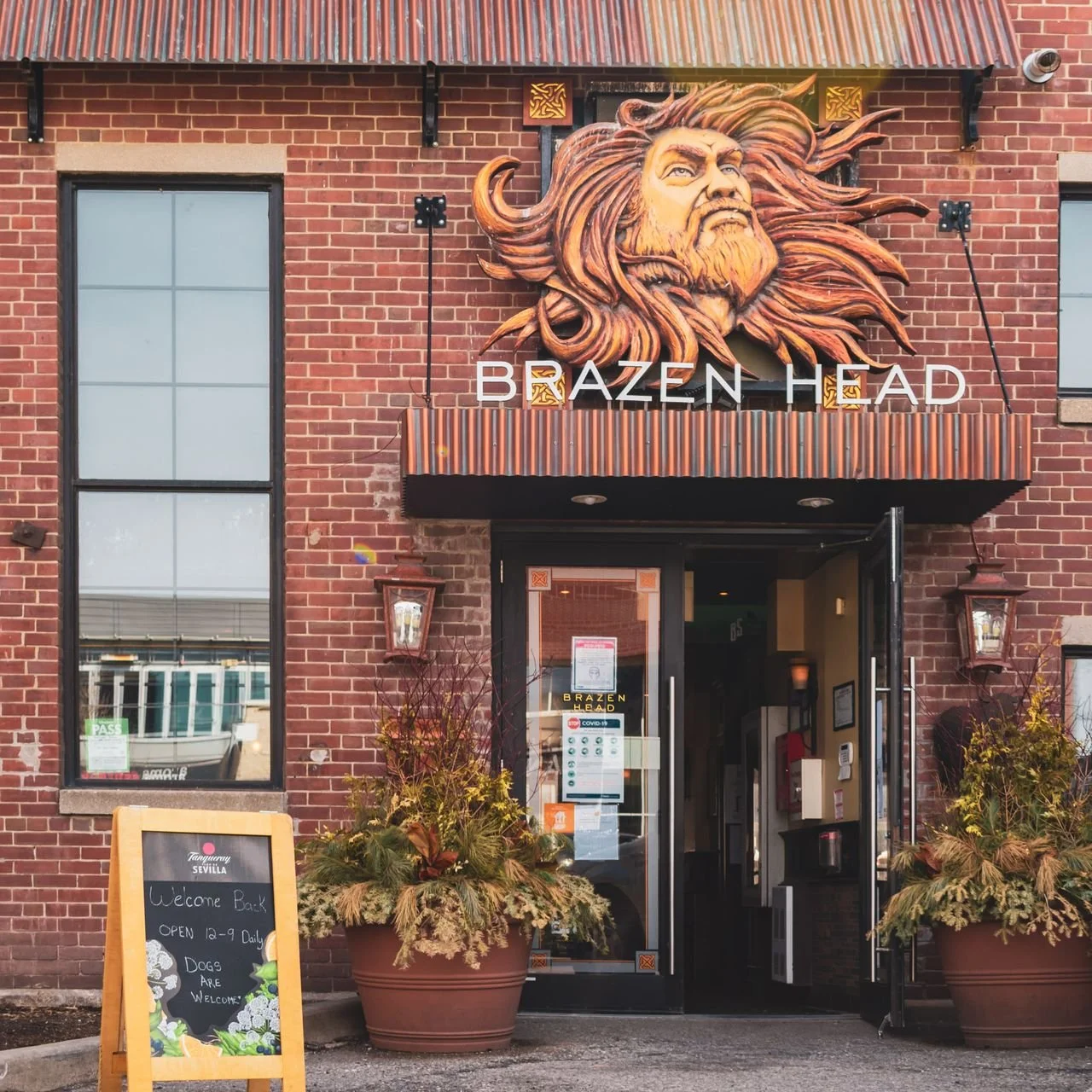 Outdoor patio seating area with tables, chairs, and umbrellas in front of a brick building with a sign for Brazen Head, metal patio furniture, and greenery