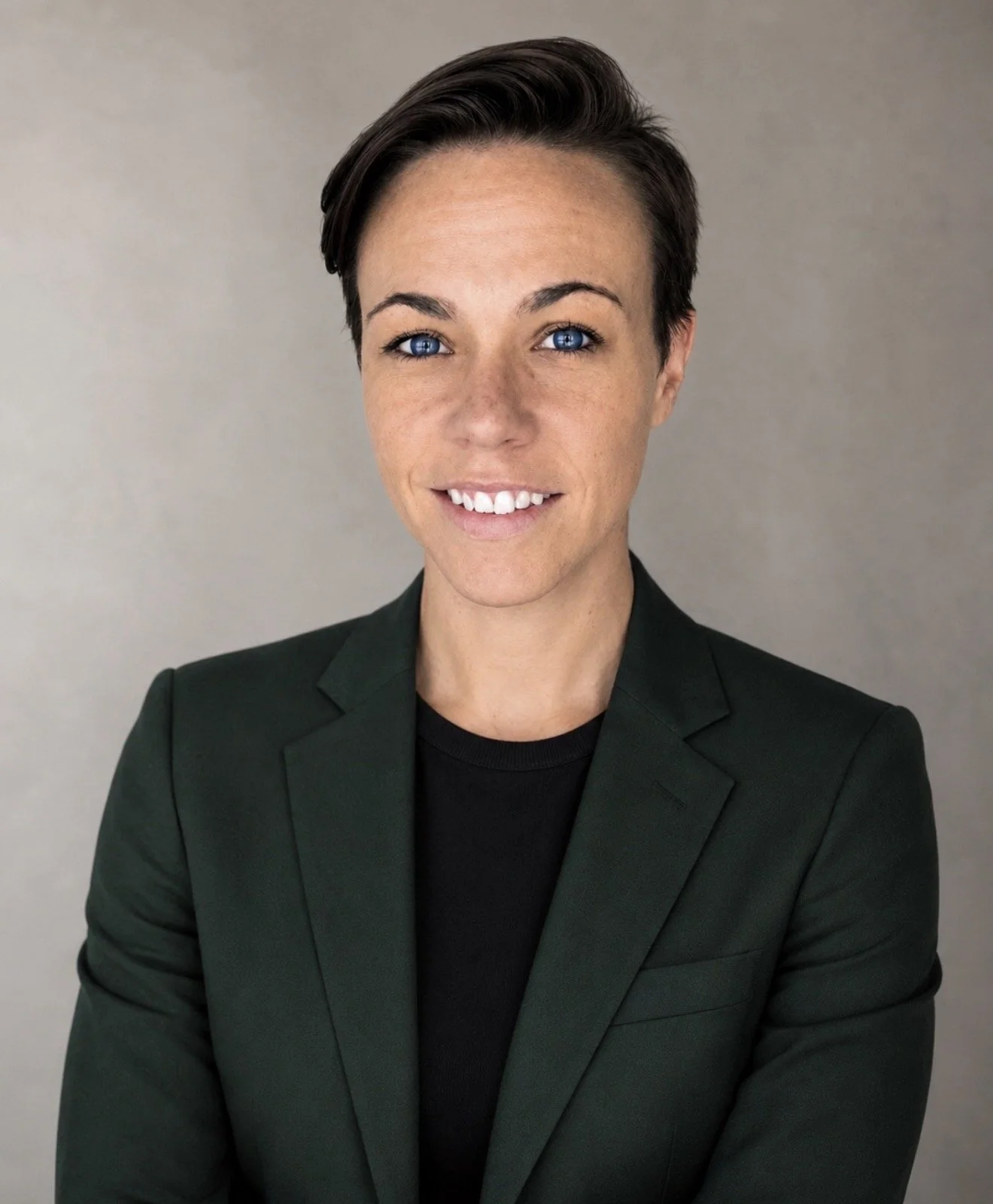 A woman with short dark hair, blue eyes, and a light complexion, smiling and wearing a black top and a dark green blazer, against a plain light background.