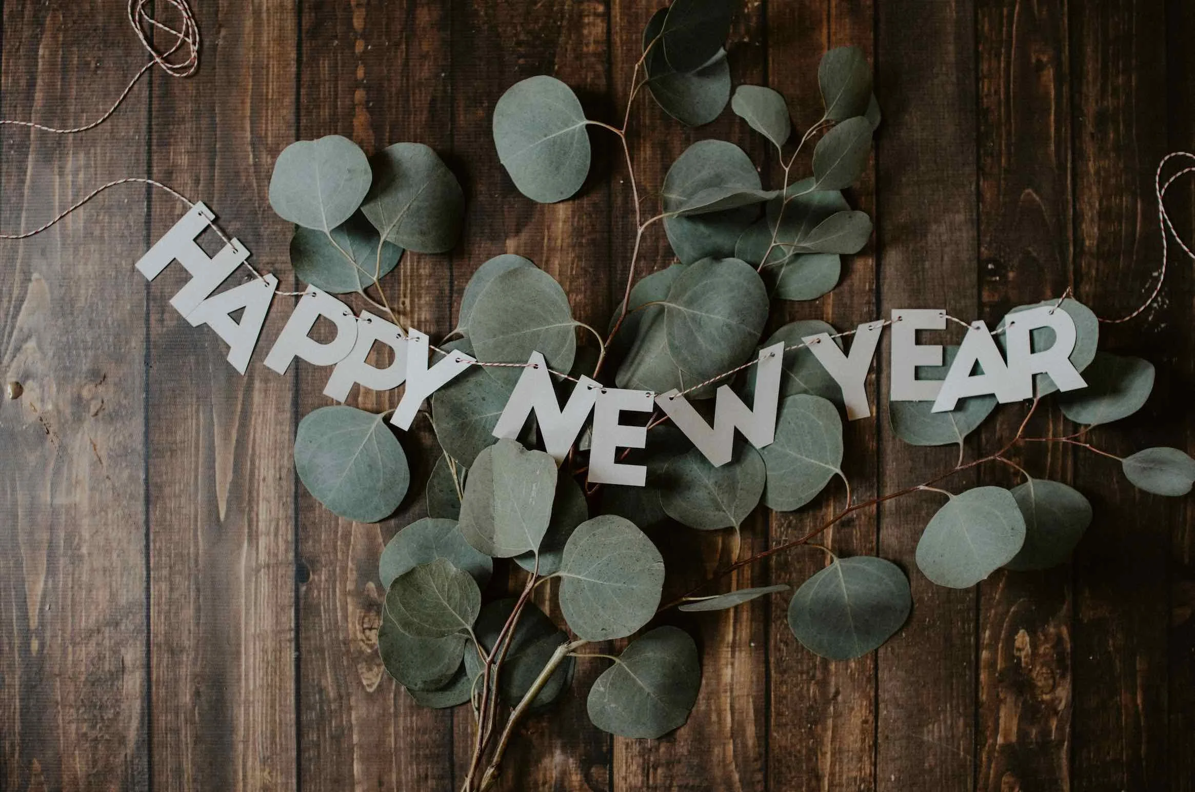 A wooden surface decorated with a string of white letters spelling 'HAPPY NEW YEAR' and eucalyptus leaves.