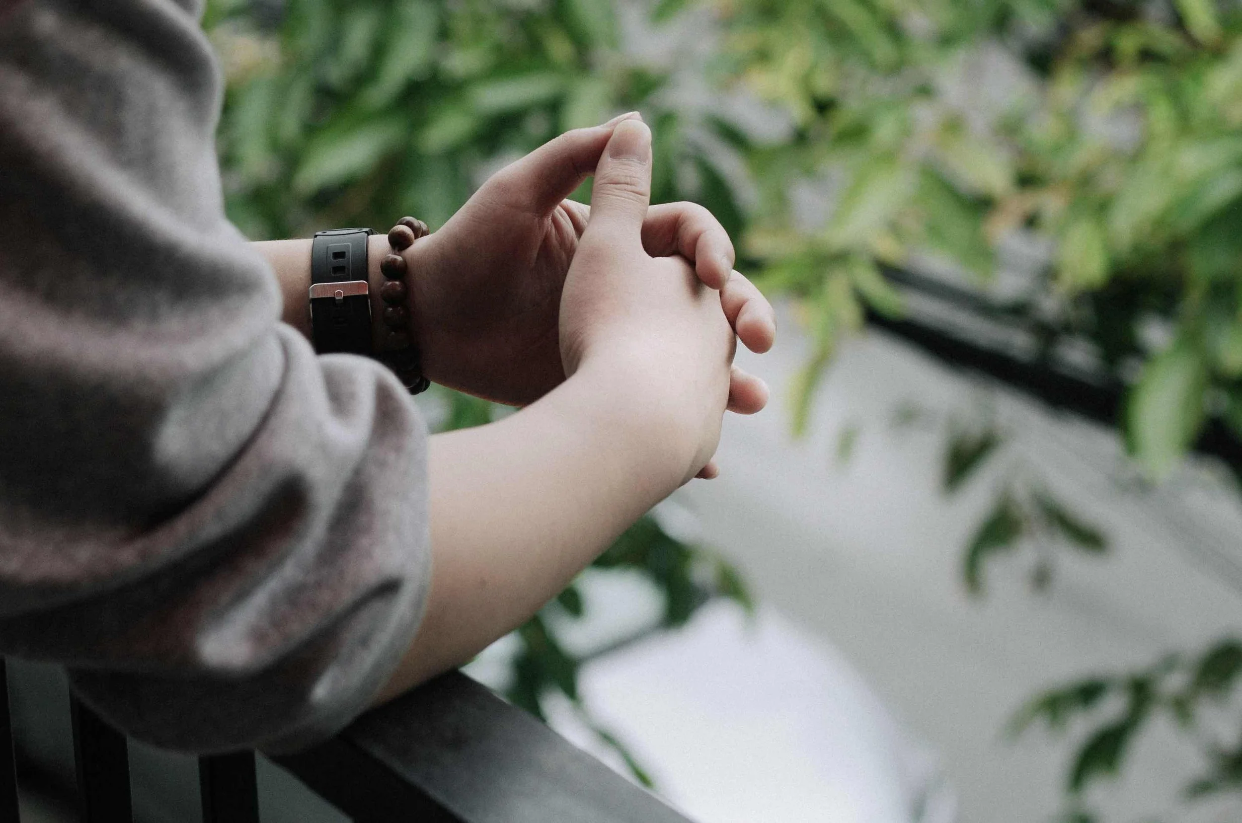 Person sitting with hands pressed together, wearing a gray long-sleeved shirt, a black watch, and a beaded bracelet, with a leafy green plant in the background.