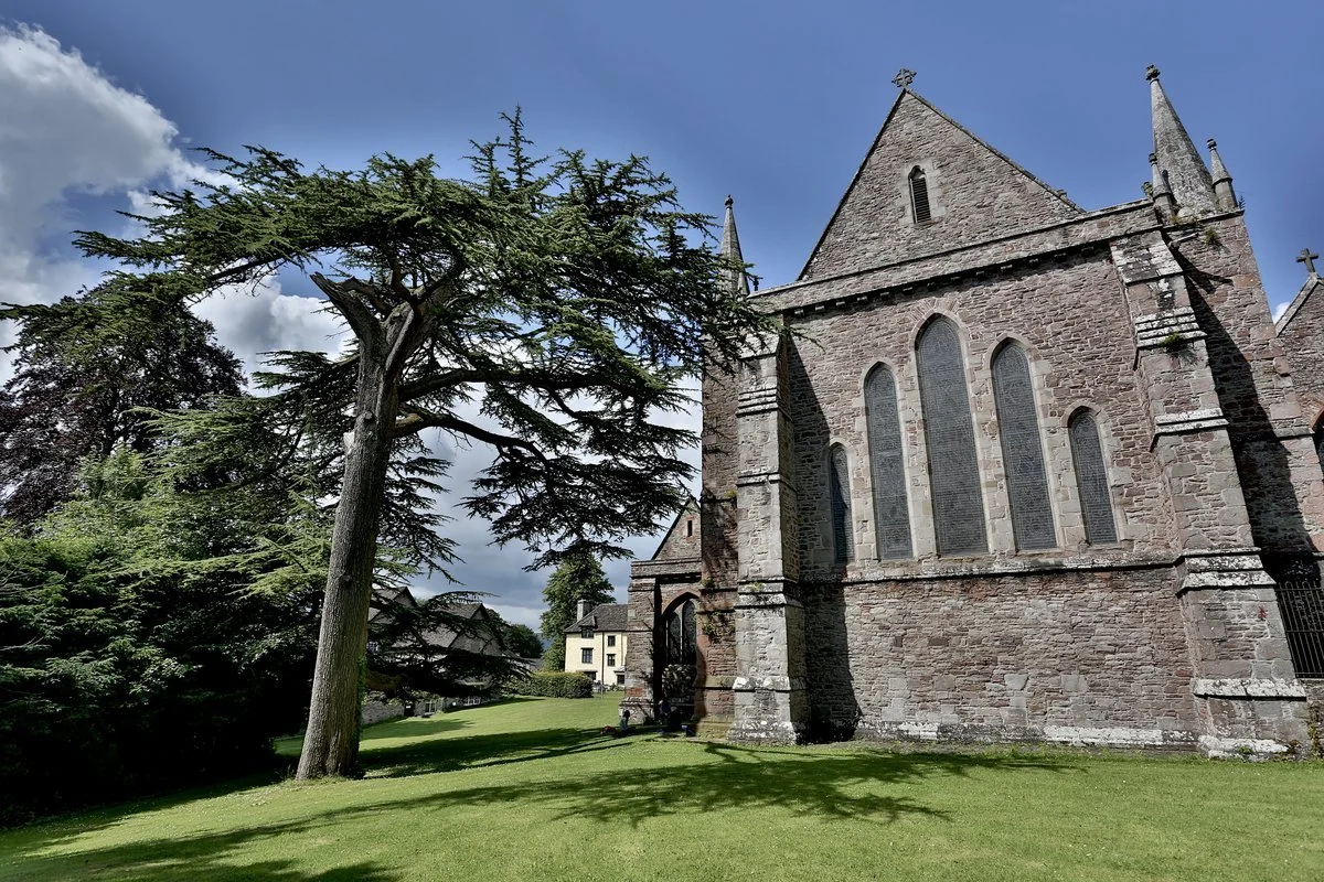 Stone church with arched windows next to a tall tree on a sunny day with blue sky and clouds.