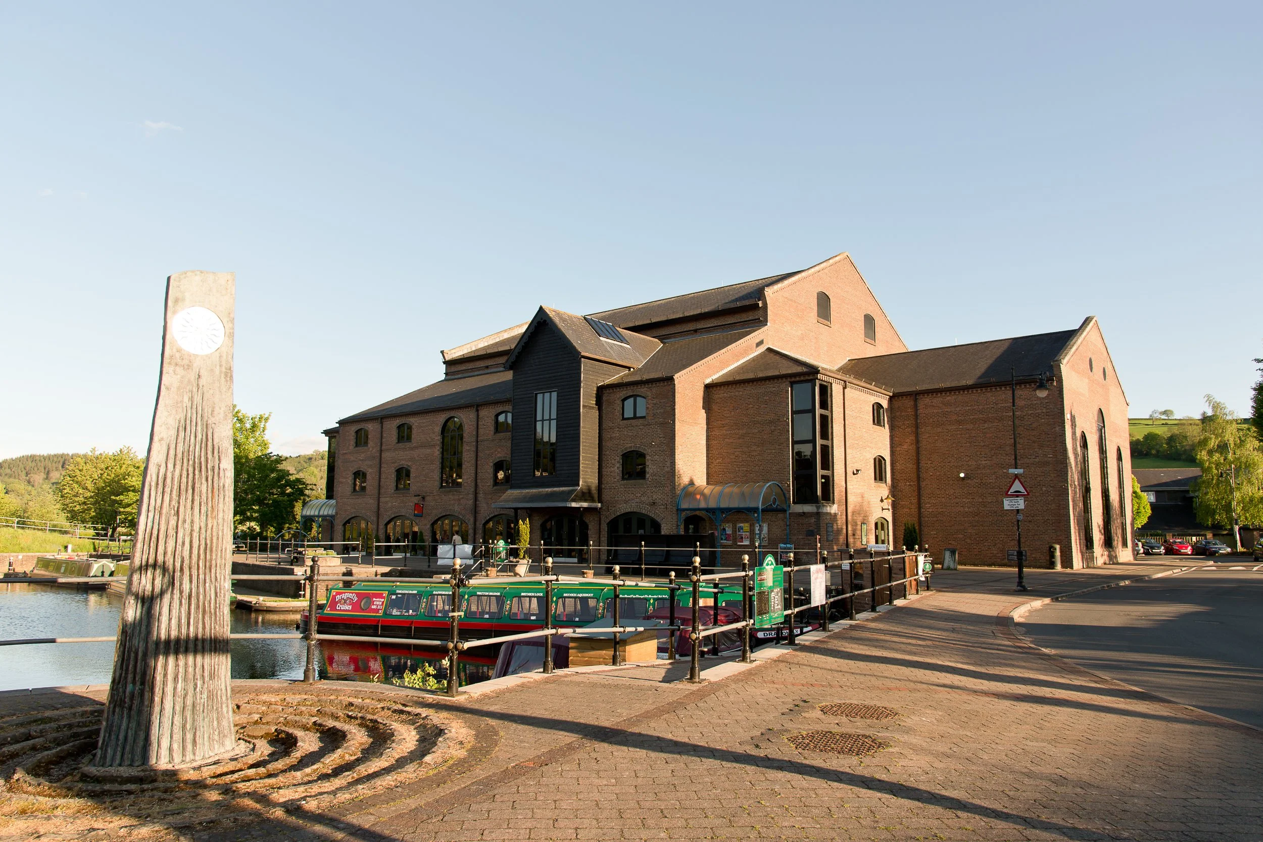 Historic brick building by a canal with narrowboat, outdoor sculpture, and clear sky.