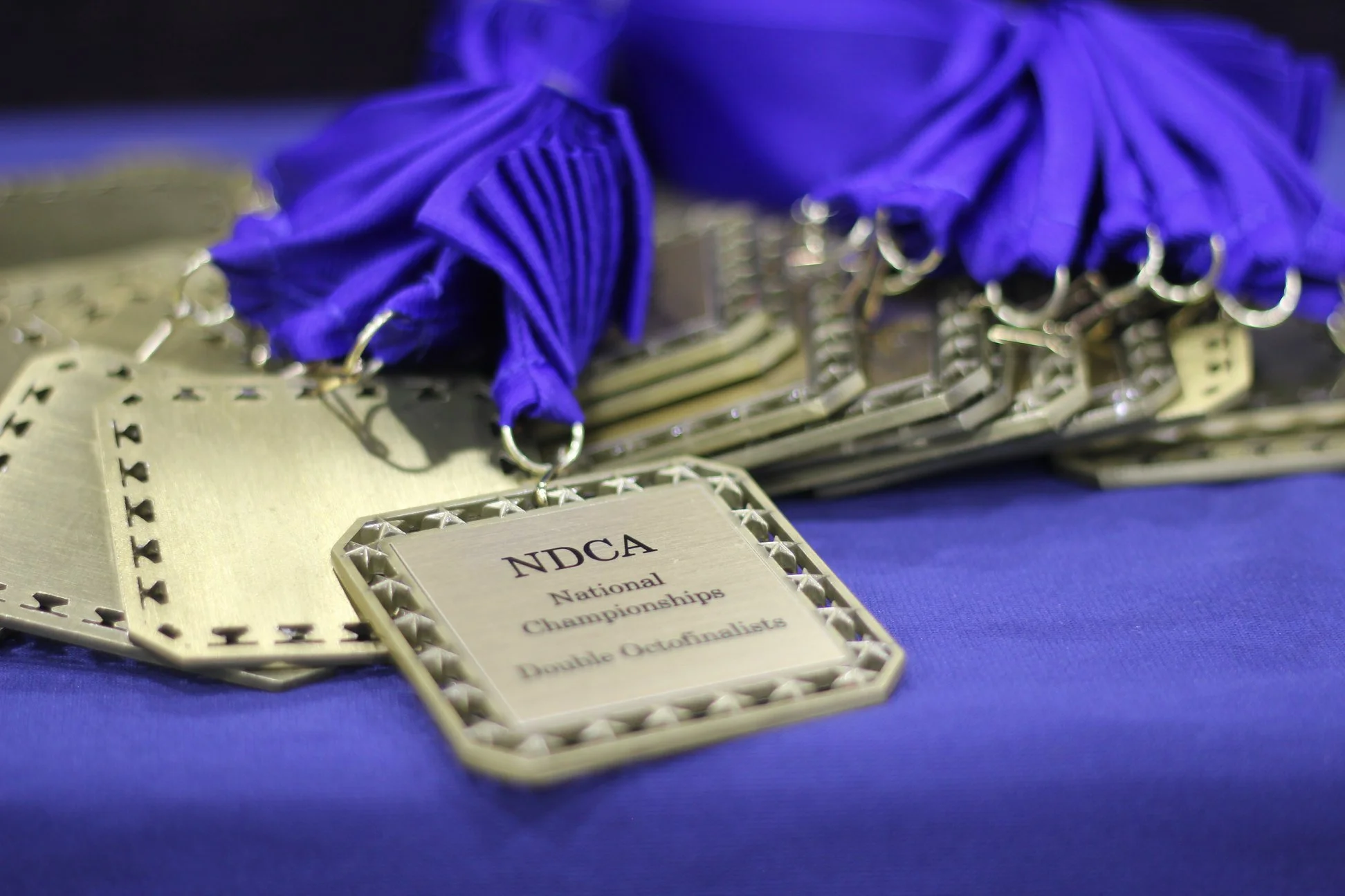 Close-up of multiple metallic championship medals with purple ribbons, displayed on a blue surface, with one medal showing the text 'NDCA National Championships Double Octofinalist'.