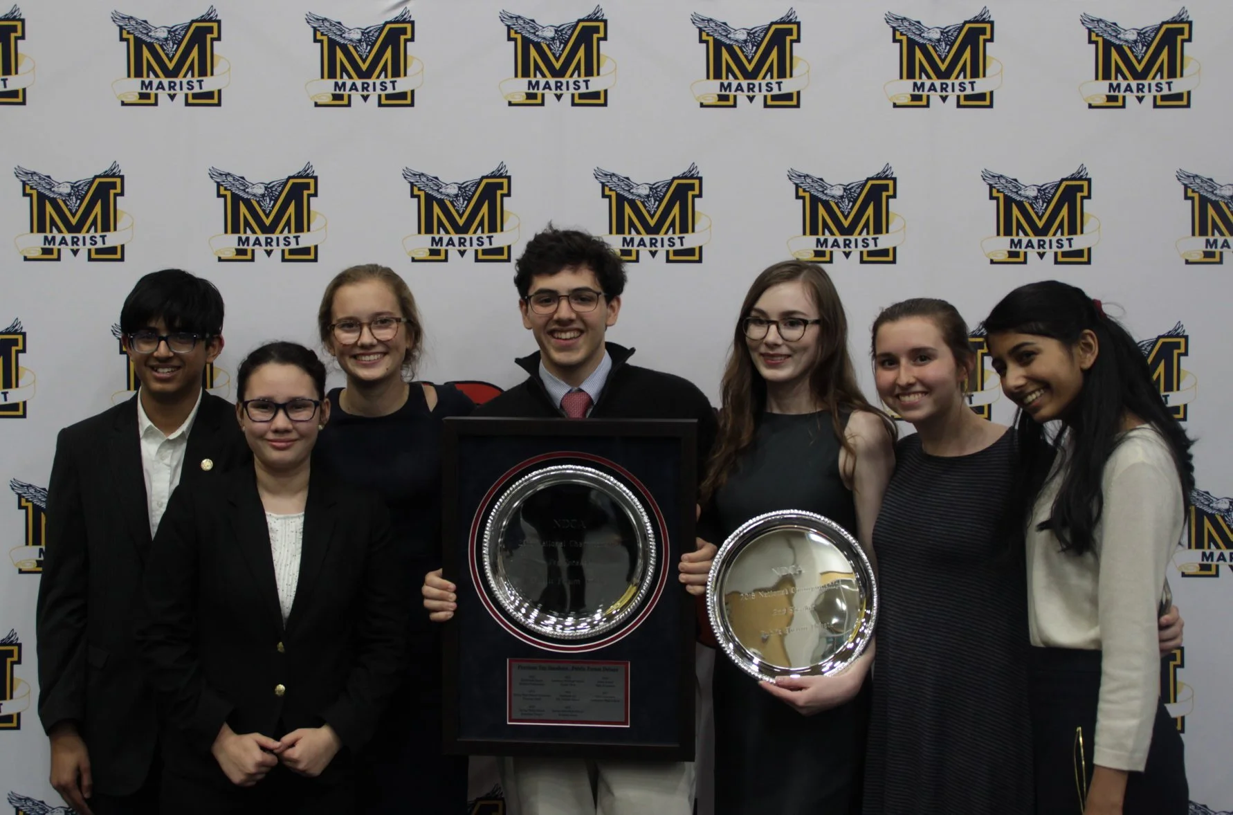 Group of six young students, three males and three females, standing together indoors in front of a school logo backdrop, holding awards after a competition or event.